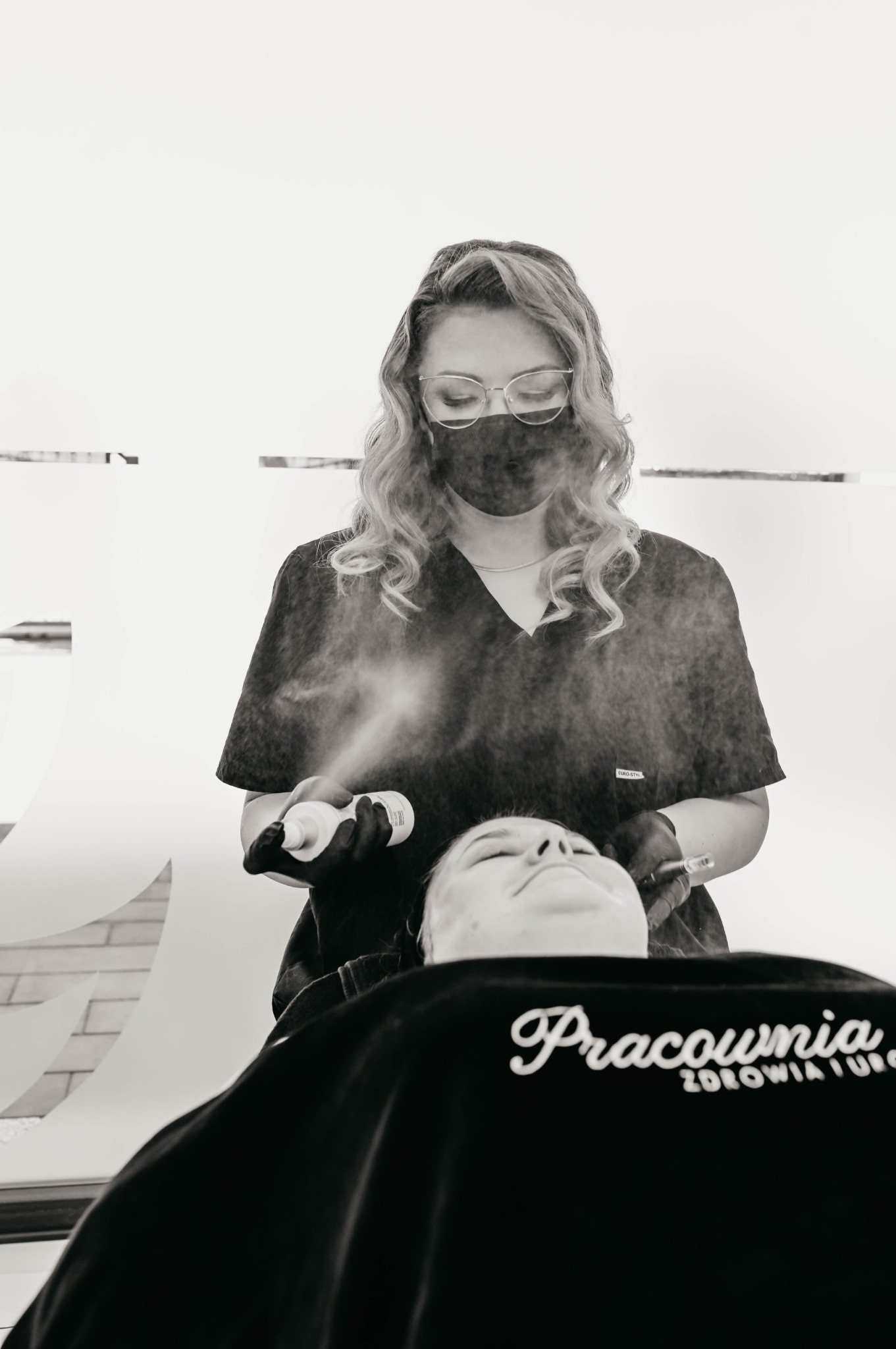 Kosmetolog w czarnym uniformie i masce spryskuje twarz klientki leżącej na leżance, przygotowując ją do zabiegu; widoczny napis 'Pracownia Zdrowia i Urody' na pelerynie ochronnej.