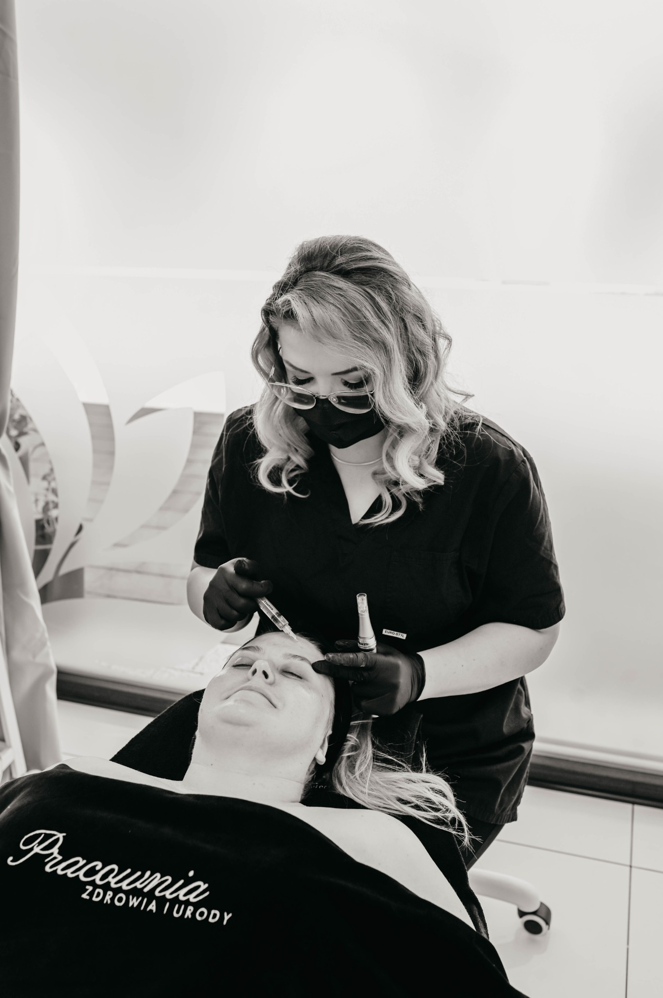 Czarno-białe zdjęcie z bliska: kosmetolog w czarnej masce i rękawiczkach wykonuje zabieg na twarzy klientki leżącej na fotelu, używając strzykawki z igłą; widoczny napis 'Pracownia Zdrowia i Urody'...