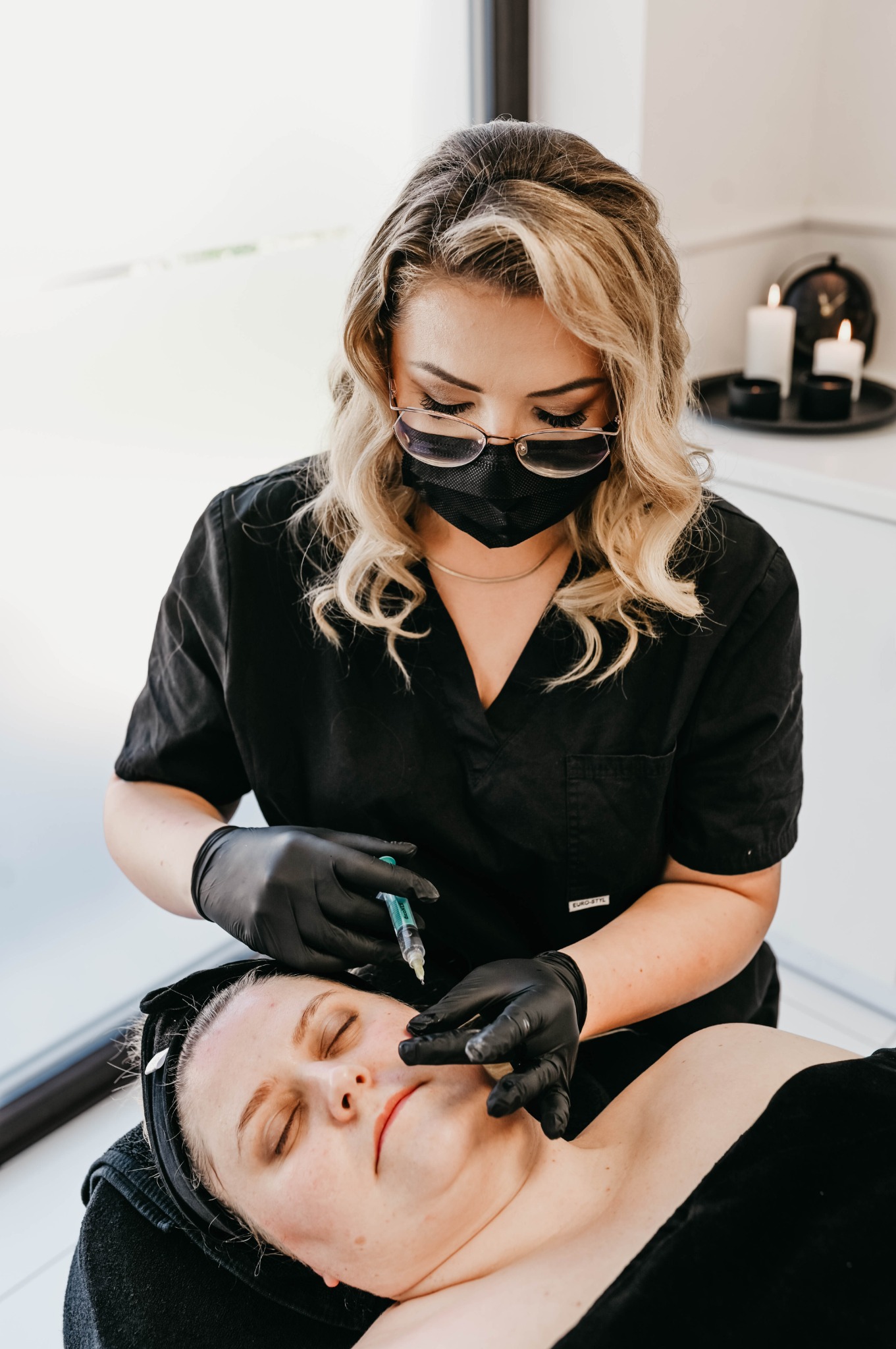 Kobieta w czarnym uniformie i rękawiczkach wykonuje iniekcję w policzek pacjentki leżącej na leżance, w tle widoczne świece i dekoracje.