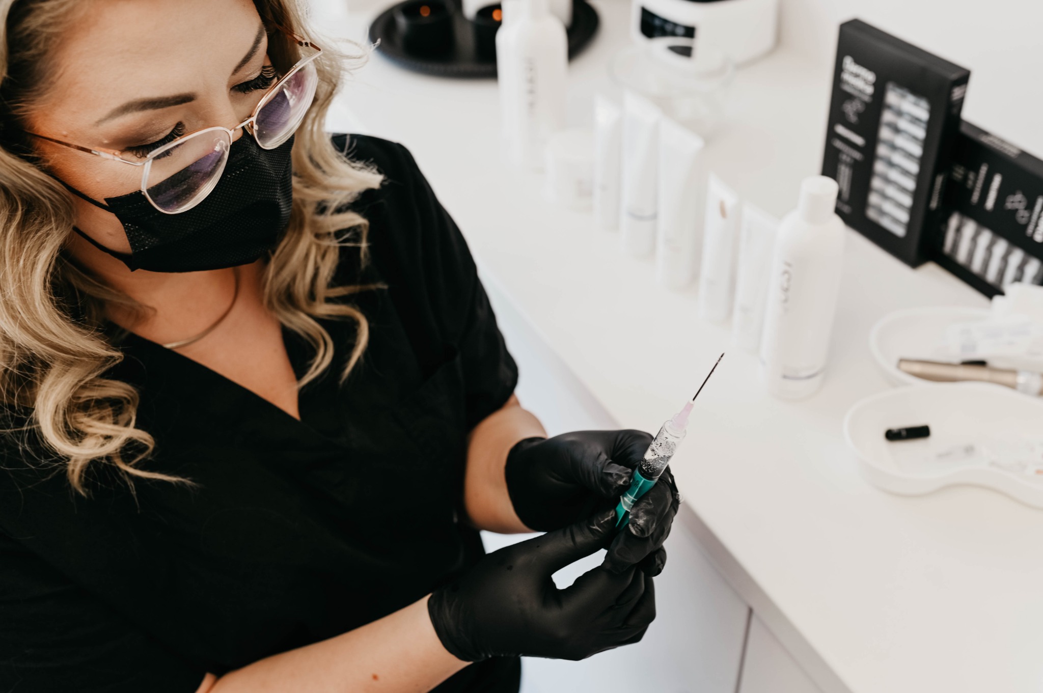 Kobieta w czarnym uniformie, okularach i maseczce, w czarnych rękawiczkach trzyma strzykawkę z zielonym płynem na tle białego blatu z kosmetykami i akcesoriami.