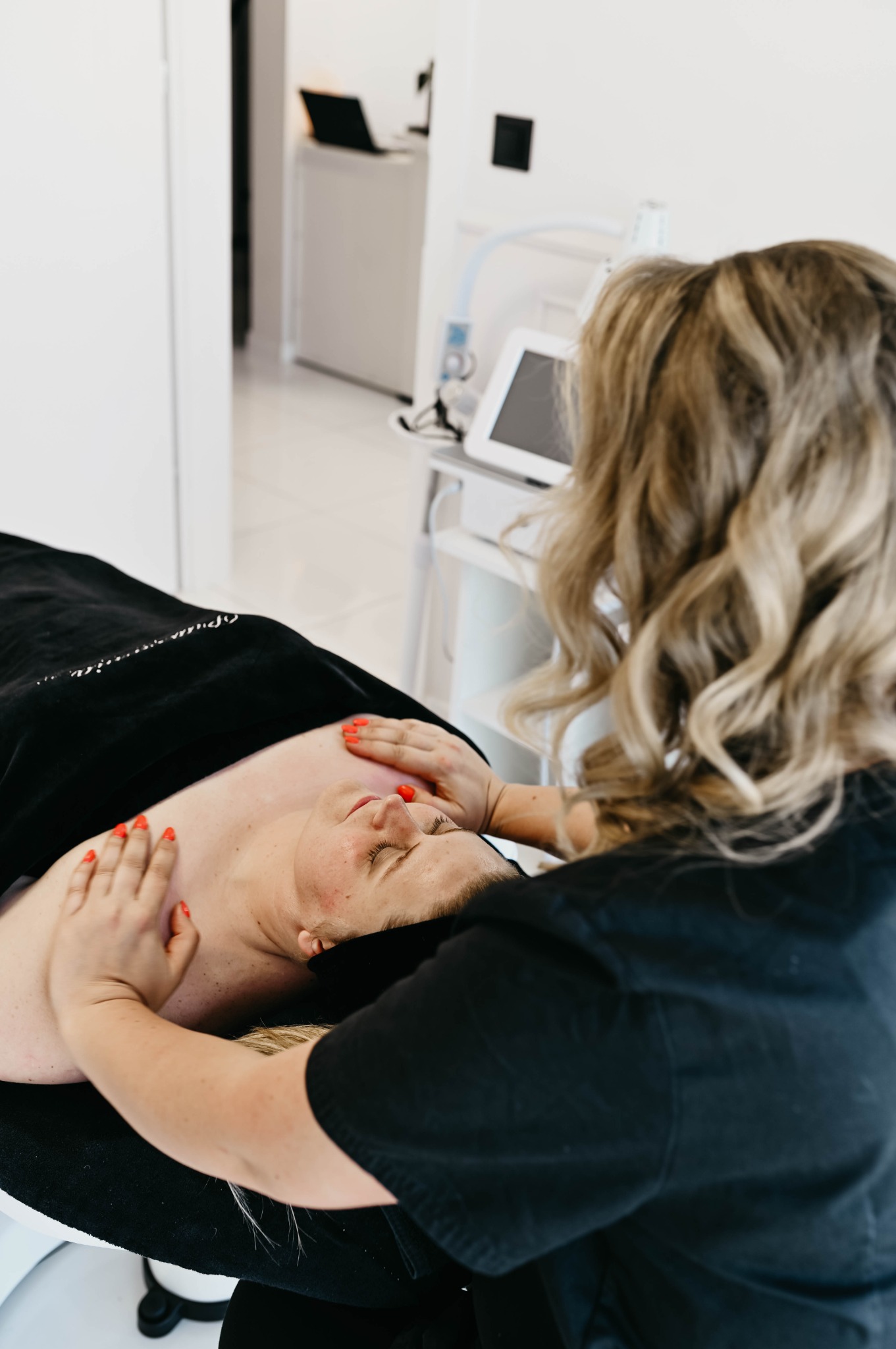 Klientka leży na leżance pod czarnym okryciem, kosmetyczka wykonuje masaż twarzy. W tle widoczny sprzęt kosmetyczny.