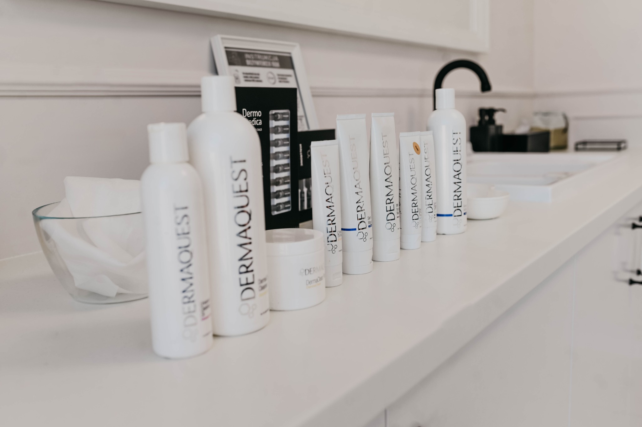 Rząd białych butelek i tubek kosmetyków marki Dermaquest ustawionych na białym blacie w salonie kosmetycznym, obok umywalki i dozownika mydła.