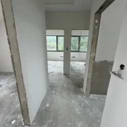 Wnętrze w trakcie remontu: surowy betonowy filar, świeżo pomalowane ściany na biało, widoczne okna z zielenią na zewnątrz, surowa podłoga z resztkami farby i gładzi, wystające kable elektryczne.