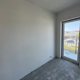 Pusty pokój z gładkimi, białymi ścianami i surową podłogą. Widoczne okno balkonowe z widokiem na zewnątrz. Minimalistyczne wnętrze w trakcie wykańczania.