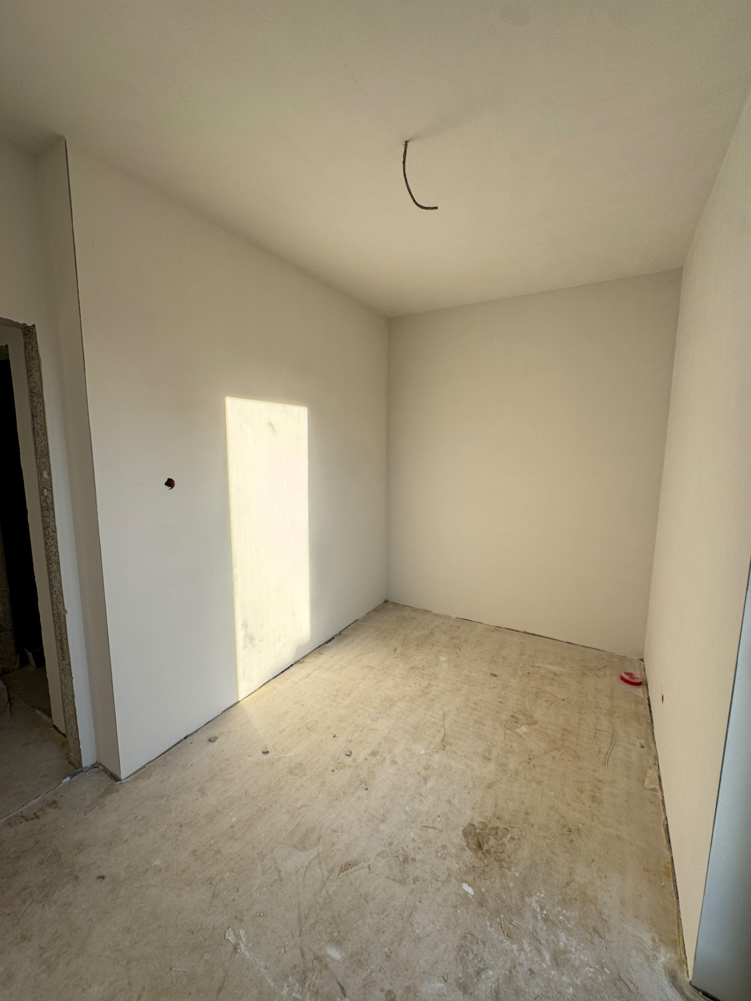 Pokój w trakcie prac wykończeniowych: świeżo zagruntowane ściany w odcieniach bieli, surowy beton na podłodze z widocznymi śladami po poprzednich pracach, wystający kabel ze ściany i światło...