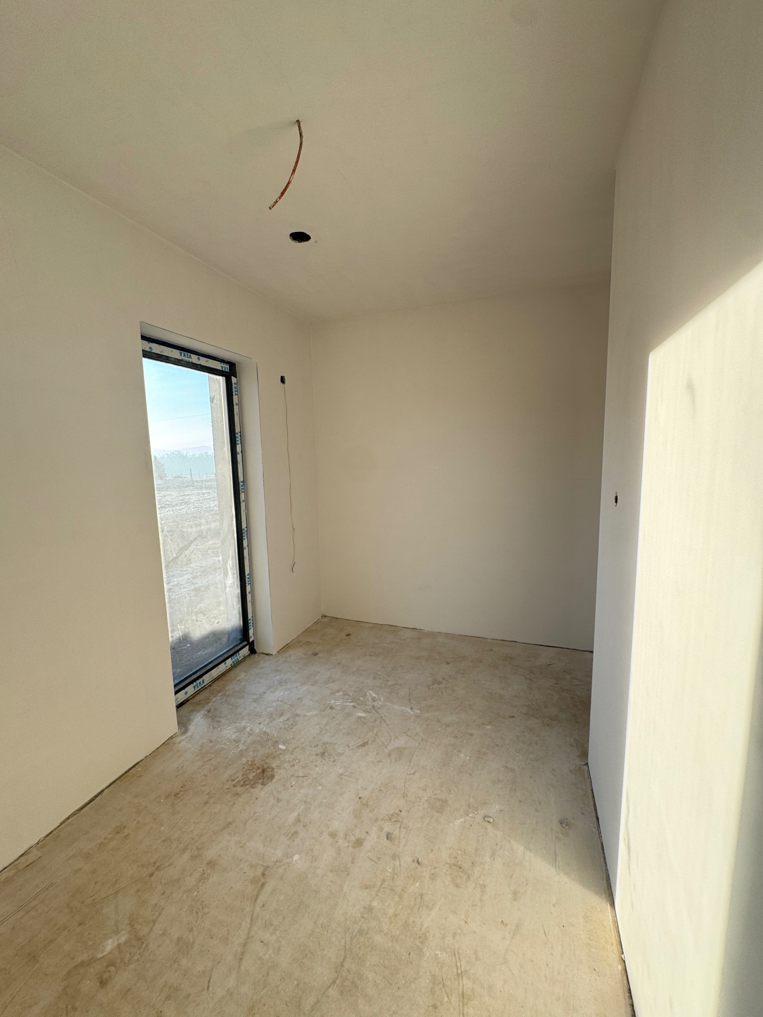 Surowe wnętrze pokoju z oknem i świeżo położonymi, jasnymi gładziami na ścianach, widoczny wystający kabel ze ściany oraz surowa podłoga przed dalszymi pracami wykończeniowymi.