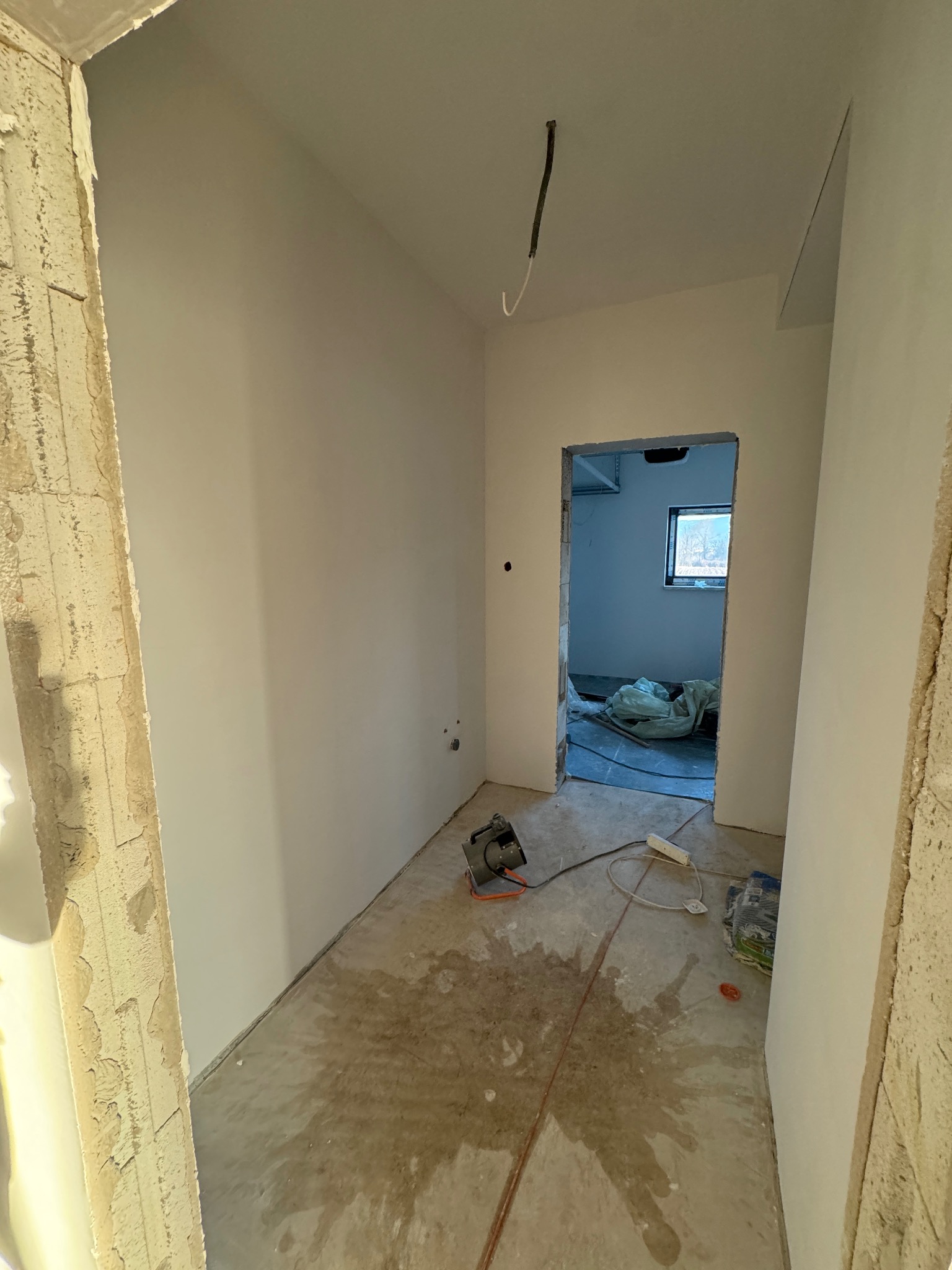 Wnętrze w trakcie wykańczania: świeżo zagruntowane ściany, surowy beton na podłodze z widocznymi śladami wilgoci, wystający kabel z sufitu i szlifirka na podłodze.