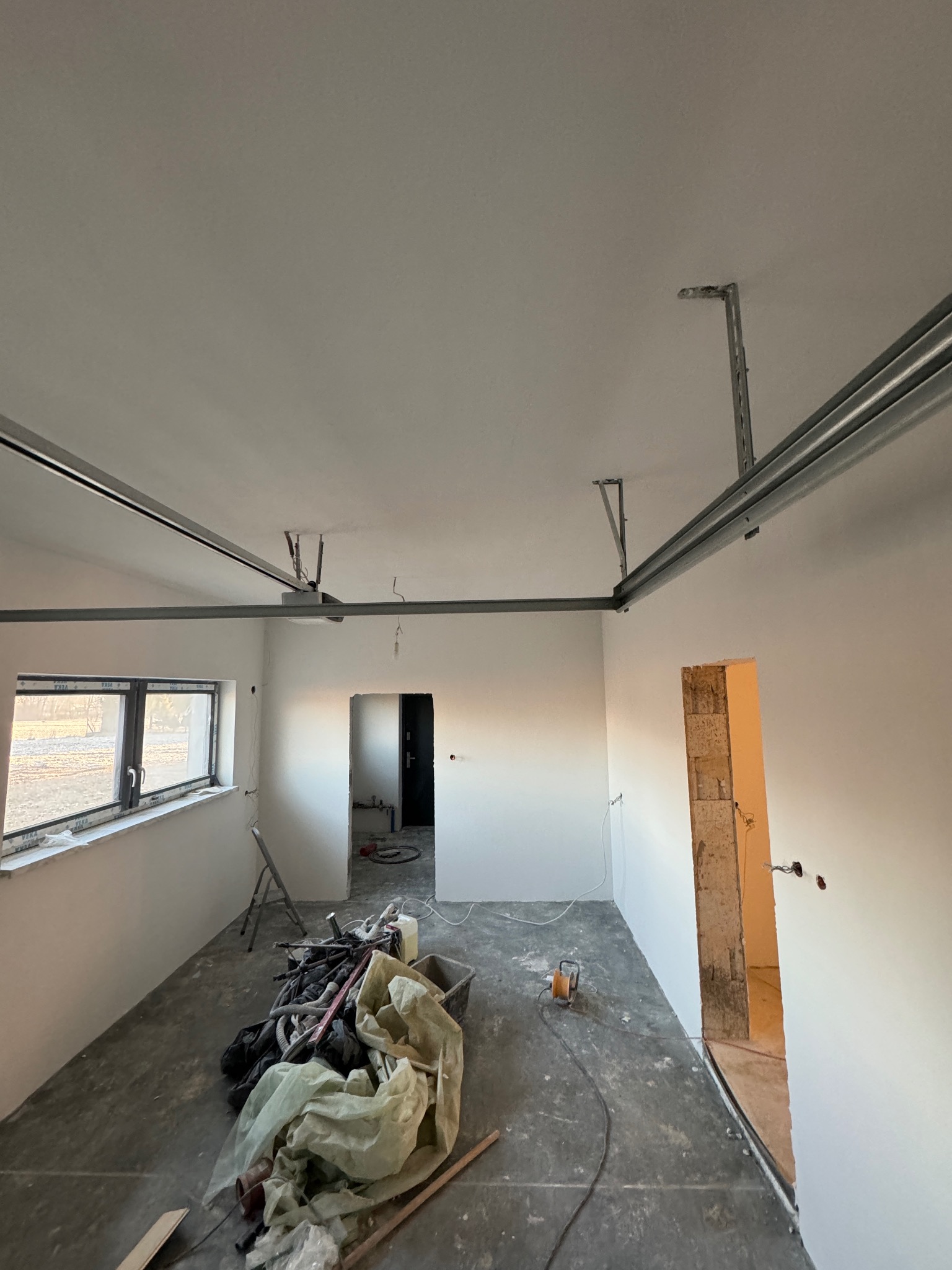 Wnętrze w trakcie prac wykończeniowych, widoczne świeżo pomalowane ściany, metalowe profile pod sufitem, przygotowane otwory drzwiowe i okienne oraz narzędzia i materiały budowlane na podłodze.