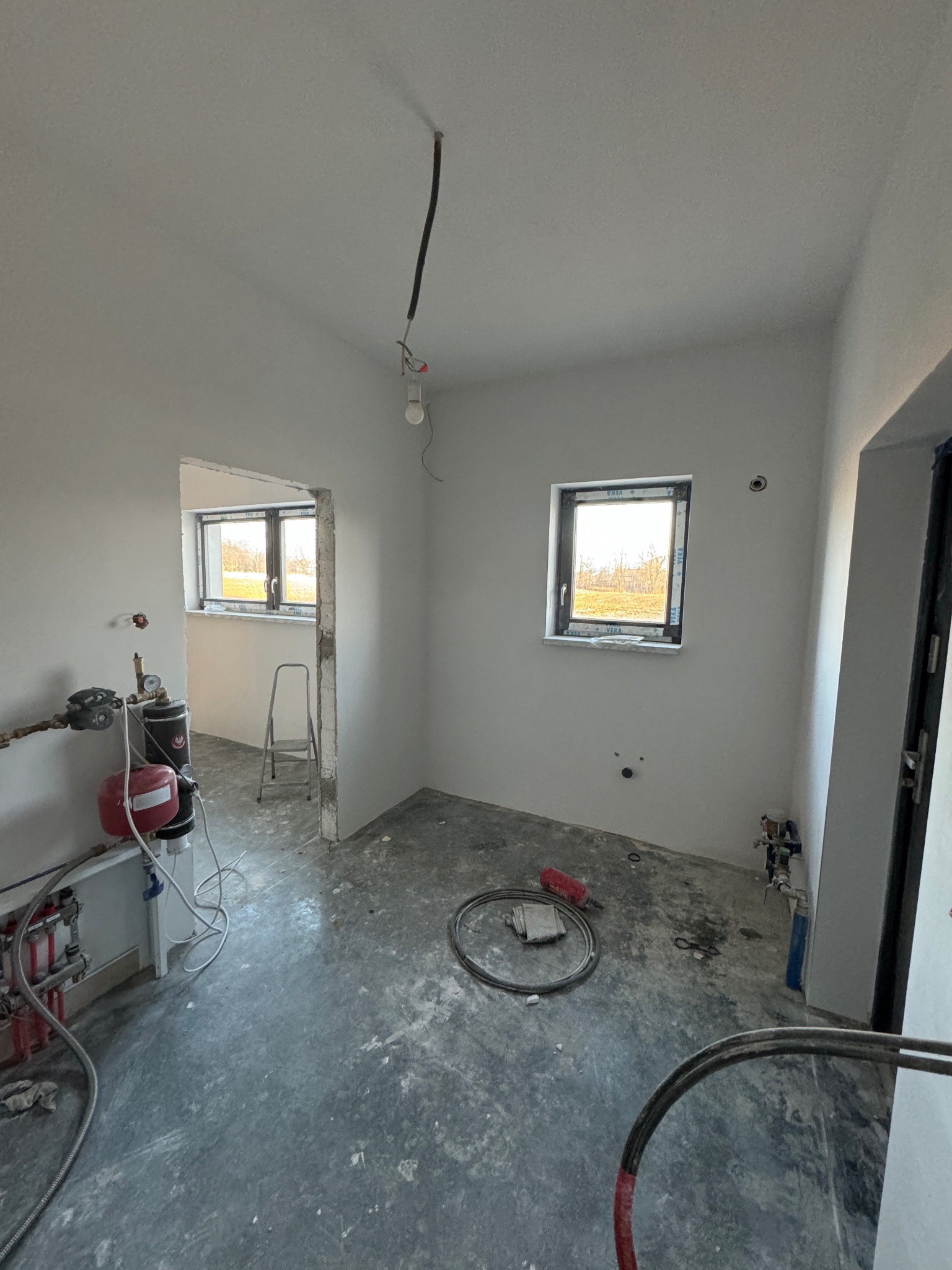 Surowe wnętrze w trakcie prac wykończeniowych: gładkie, białe ściany, betonowa podłoga, widoczne instalacje hydrauliczne, okno z widokiem na zewnątrz i wisząca żarówka na kablu.