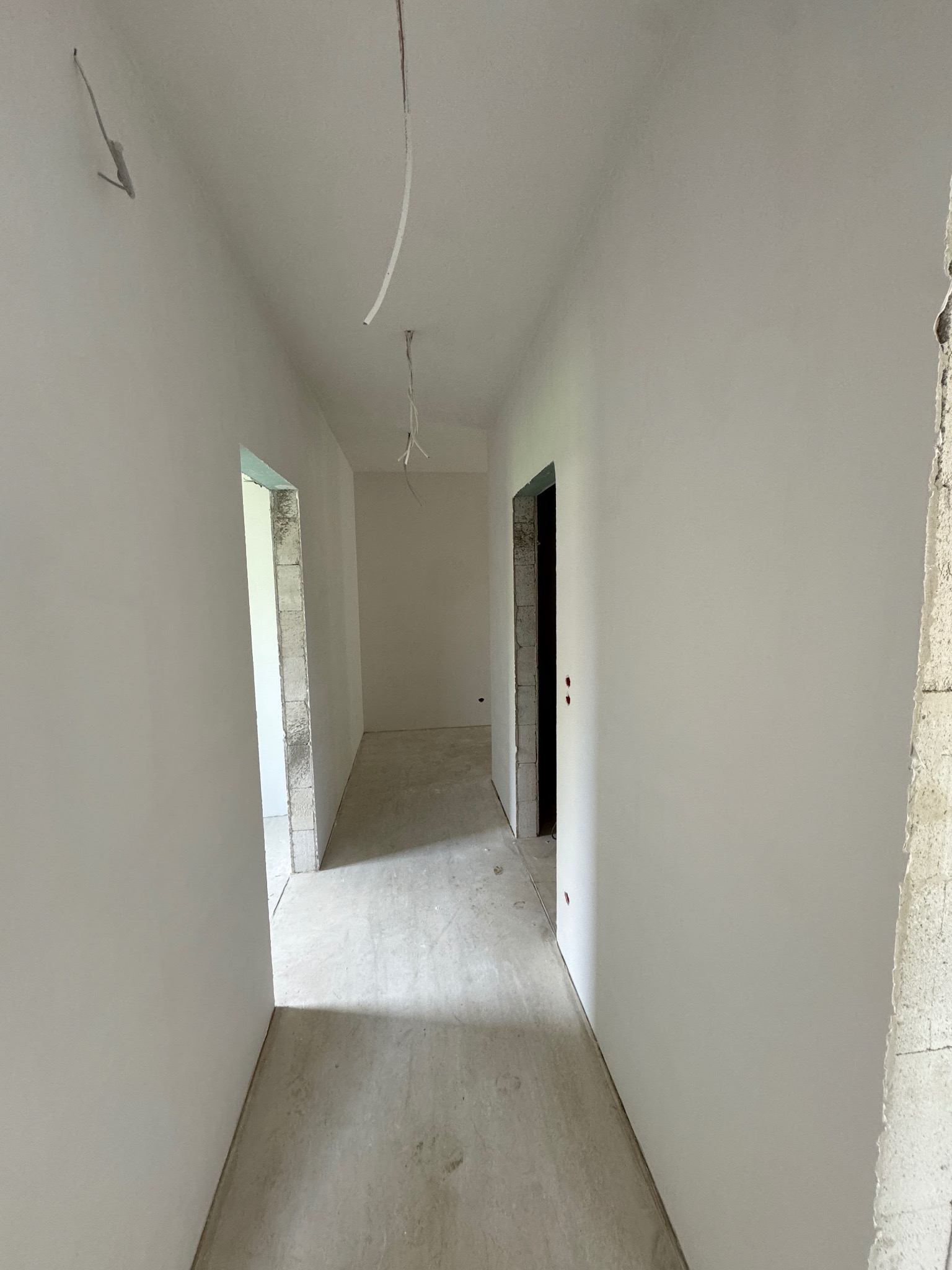 Wnętrze korytarza w trakcie prac wykończeniowych, ściany po gładziowaniu, widoczne wystające przewody elektryczne i surowy beton przy otworach drzwiowych.