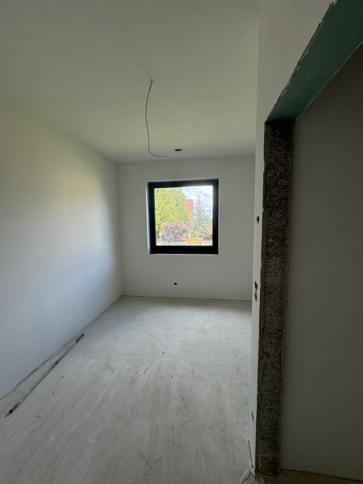 Surowe wnętrze w Pietrzykowicach: białe ściany, szare podłoże, okno z widokiem na zieleń, fragment surowego otworu drzwiowego. Widoczny kabel zwisający z sufitu.