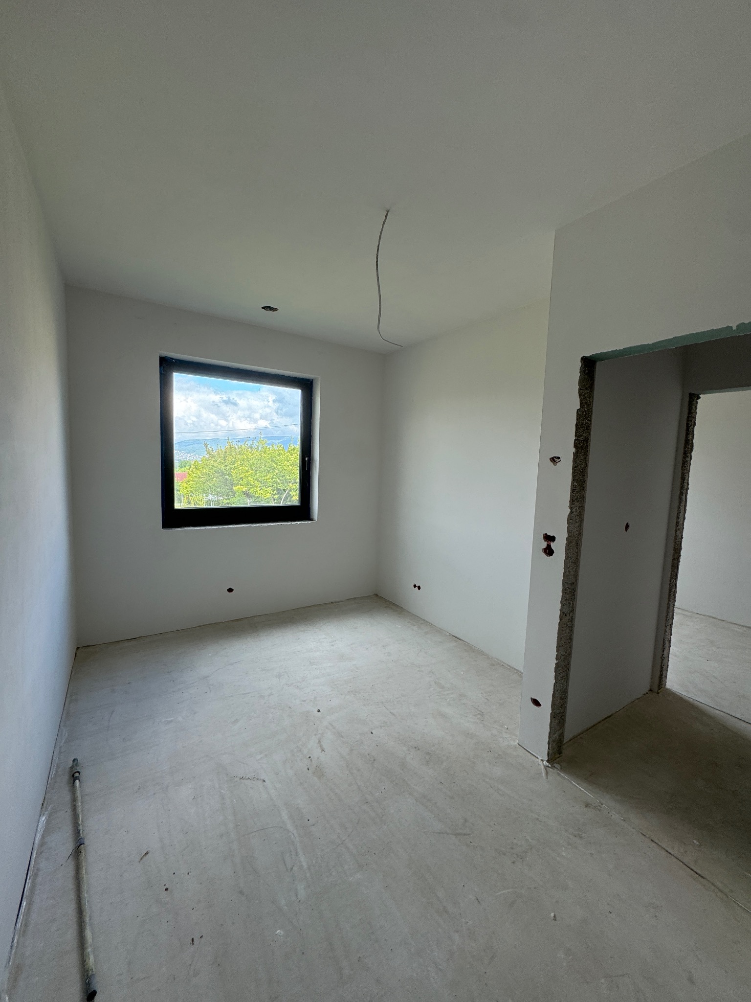 Surowe wnętrze po gładziowaniu w Pietrzykowicach: białe ściany, betonowa podłoga, czarne okno z widokiem na zieleń. Przygotowanie do dalszych prac.
