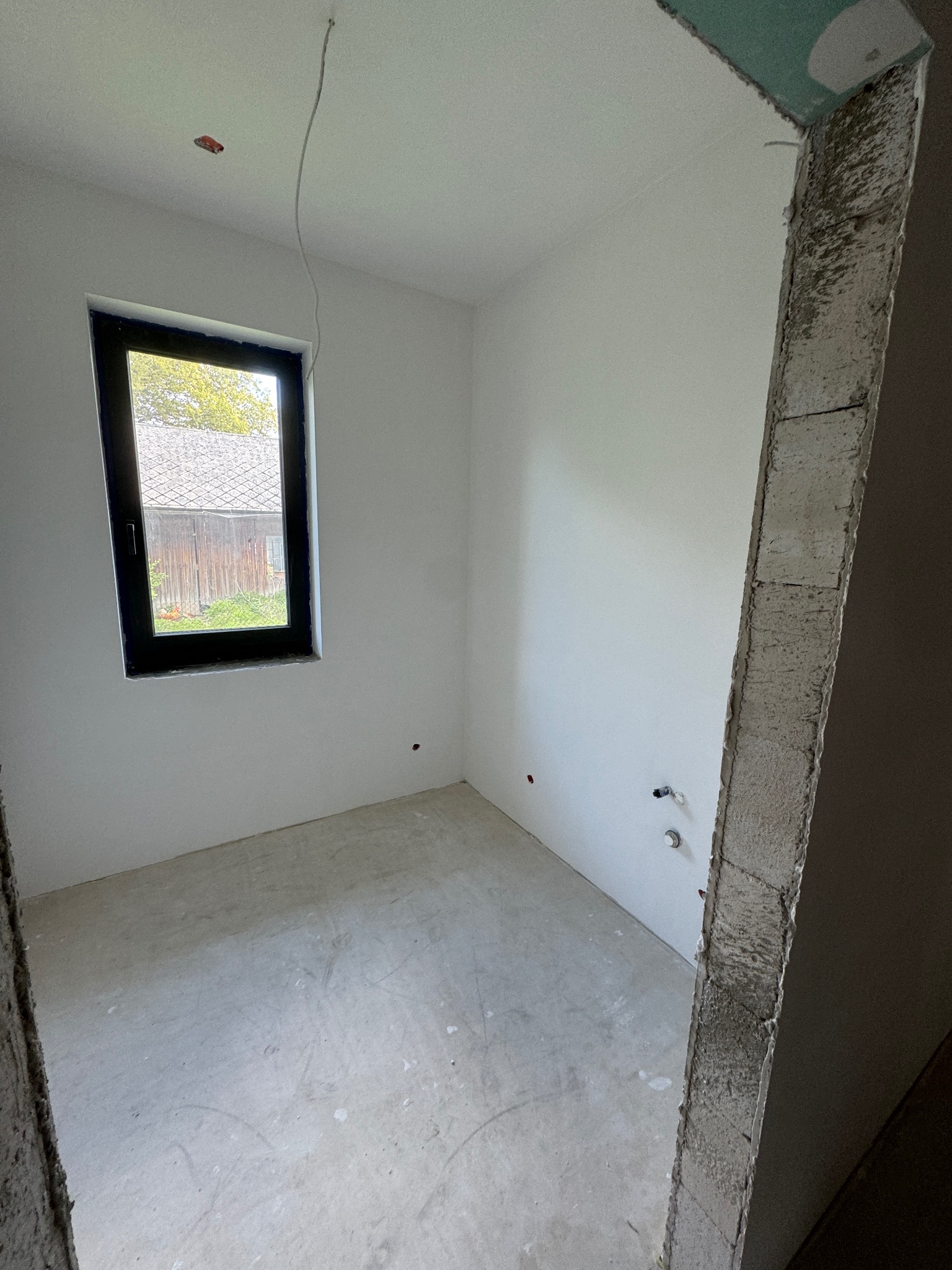 Surowe wnętrze w Pietrzykowicach: gładkie, białe ściany, czarna rama okna z widokiem na budynek gospodarczy, surowy beton na podłodze i ościeżnicy.