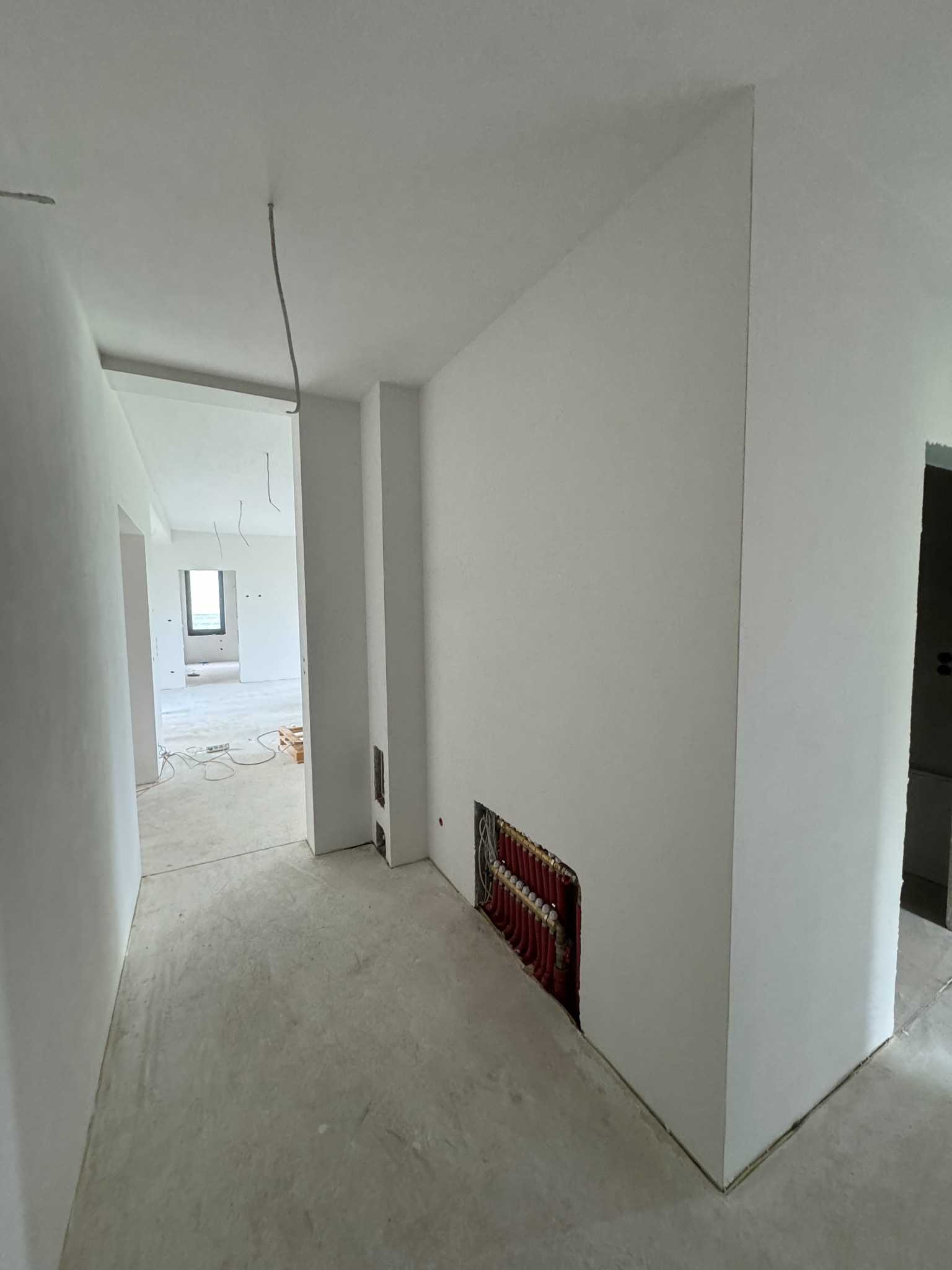 Wnętrze w trakcie prac wykończeniowych: gładkie, białe ściany, surowa podłoga, widoczne elementy instalacji ogrzewania podłogowego w ścianie i wystające kable.