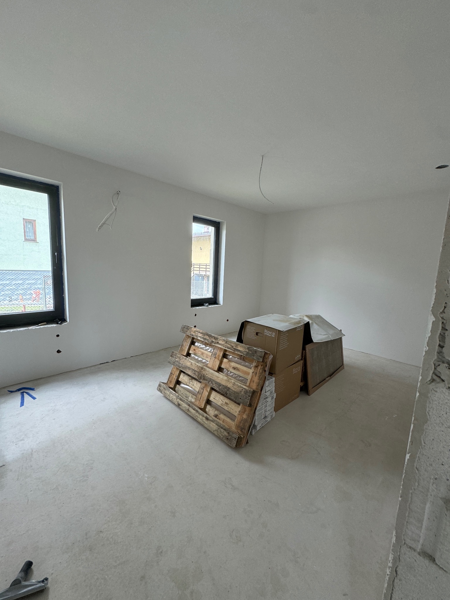 Wnętrze w trakcie wykończenia: gładkie, białe ściany, okna z czarnymi ramami, paleta, kartony i narzędzie na podłodze. W tle widok z okna. Pietrzykowice.