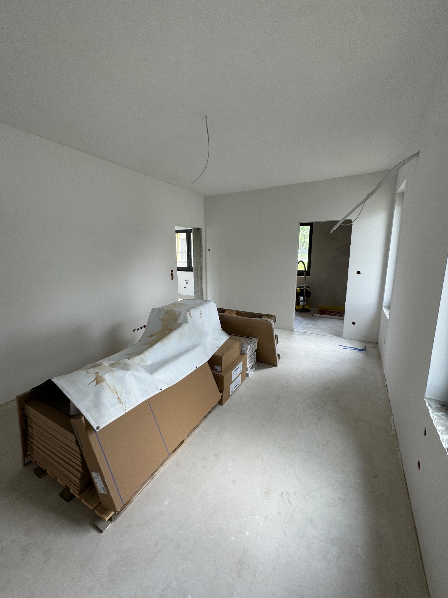 Wnętrze w trakcie prac wykończeniowych: białe ściany, podłoga wylewka, paleta z panelami pod przykryciem. Widoczne kable, otwory okienne i drzwiowe.