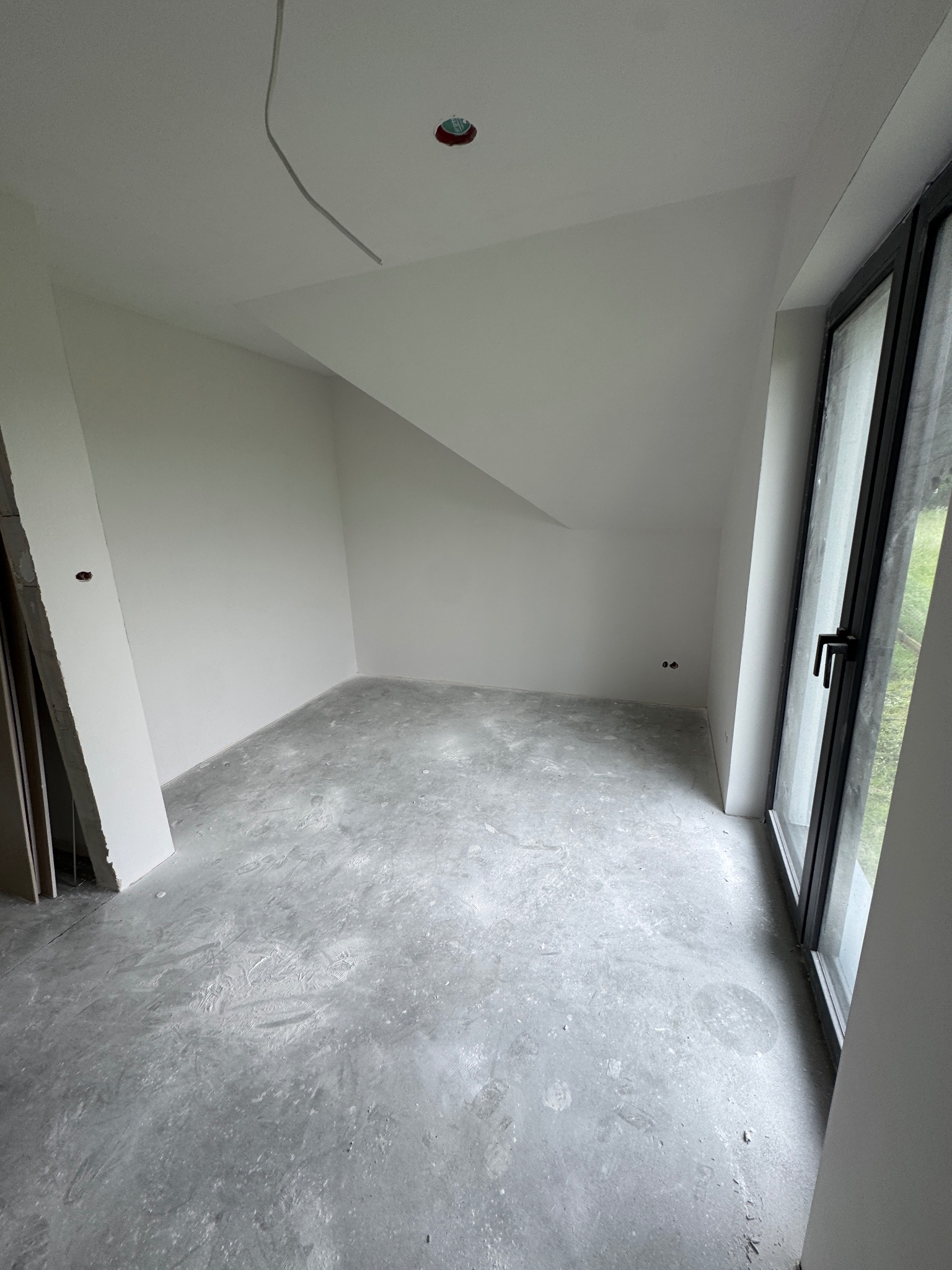 Surowe wnętrze poddasza w Radziechowach: białe ściany, szary beton na podłodze, czarne ramy okienne, widoczne instalacje elektryczne pod sufitem.