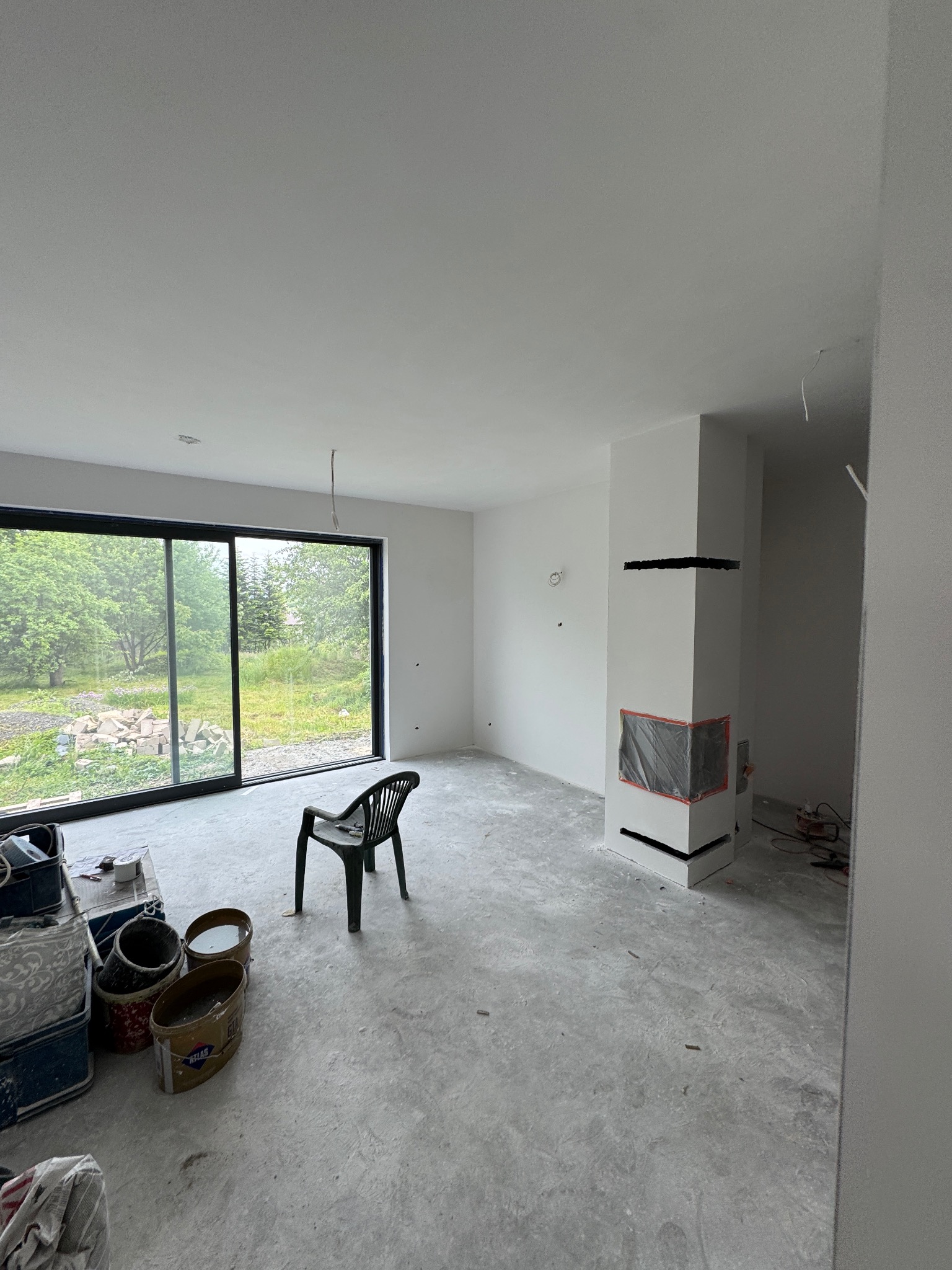 Wnętrze w Radziechowach w trakcie prac wykończeniowych: świeżo pomalowane ściany, zabezpieczony kominek, okno z widokiem na zieleń, wiadra z farbą.
