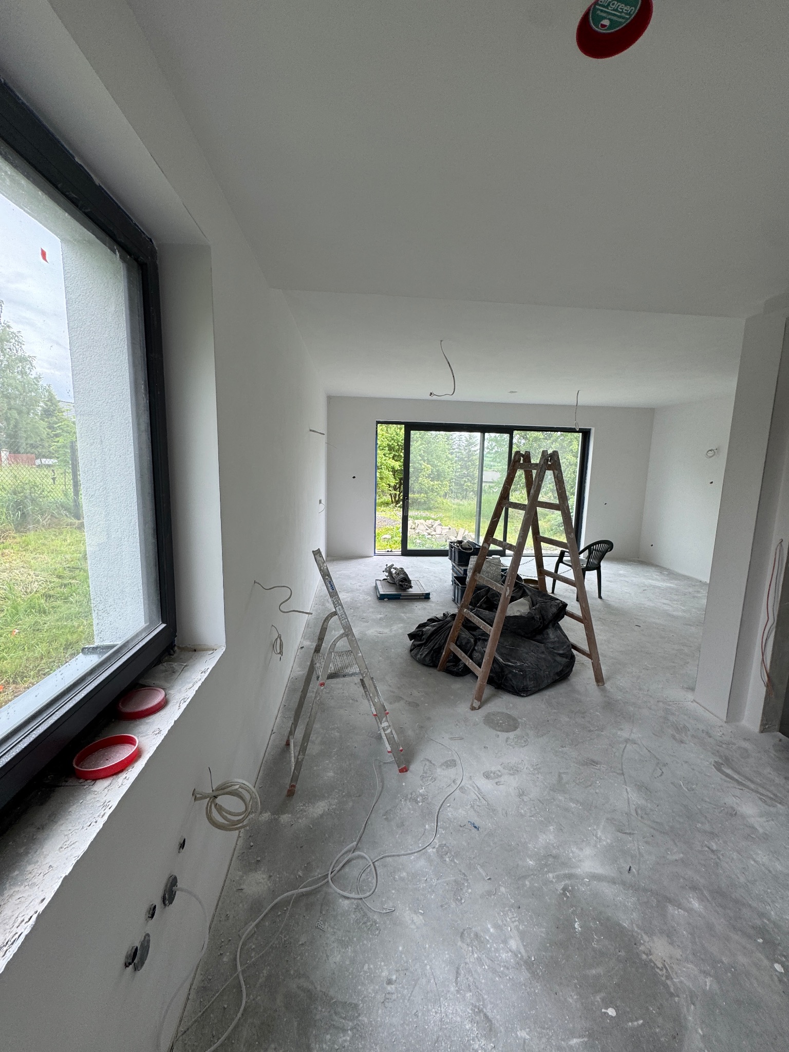 Wnętrze w trakcie prac wykończeniowych: ściany po gładziowaniu, widoczne drabiny, przewody elektryczne i okno z widokiem na zieleń. Radziechowy.