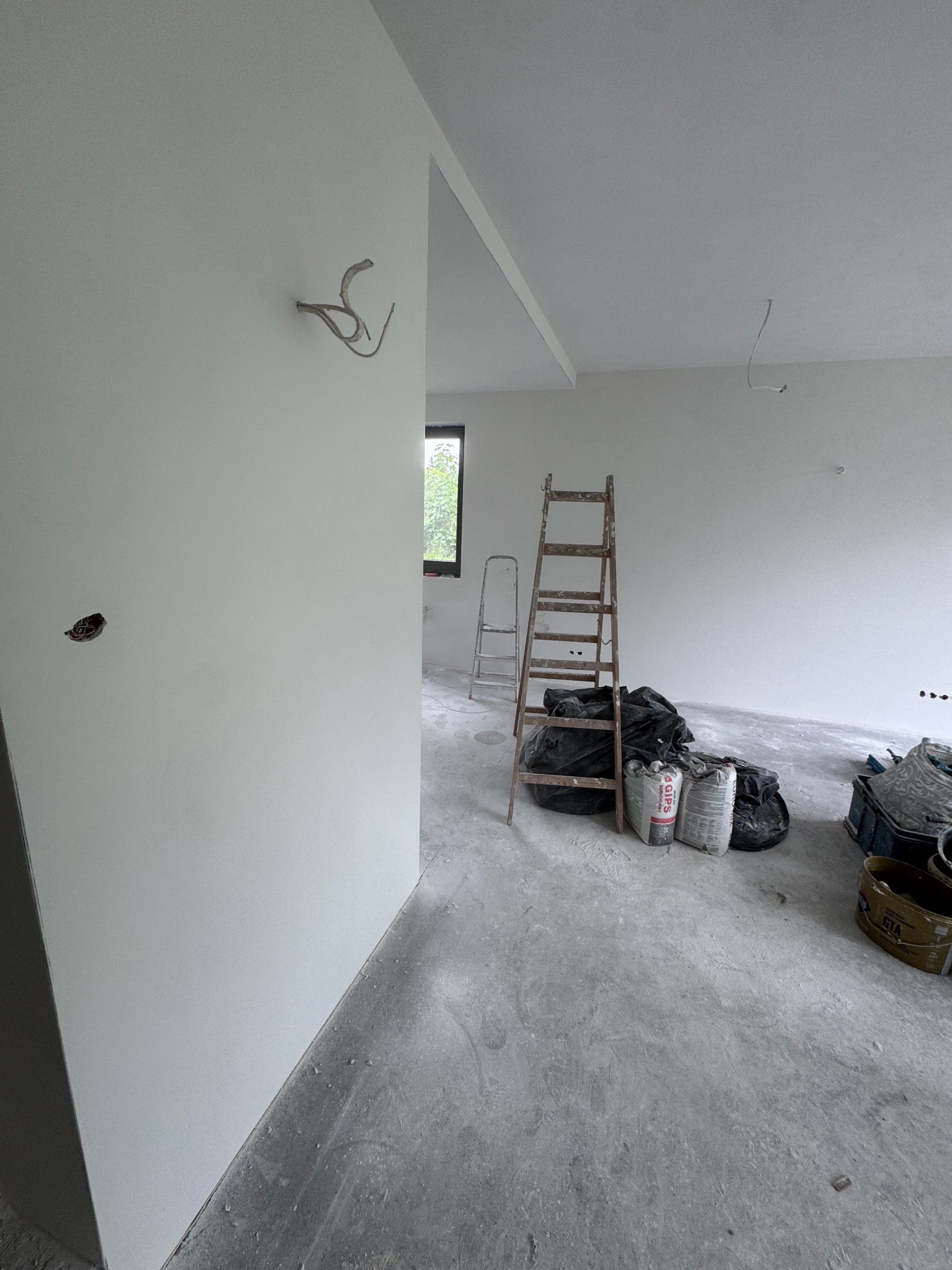 Radziechowy: Wnętrze w trakcie gładzenia ścian, widoczne worki z gipsem, drabina i wystające kable. Świeżo zagruntowane ściany i szary beton na podłodze.