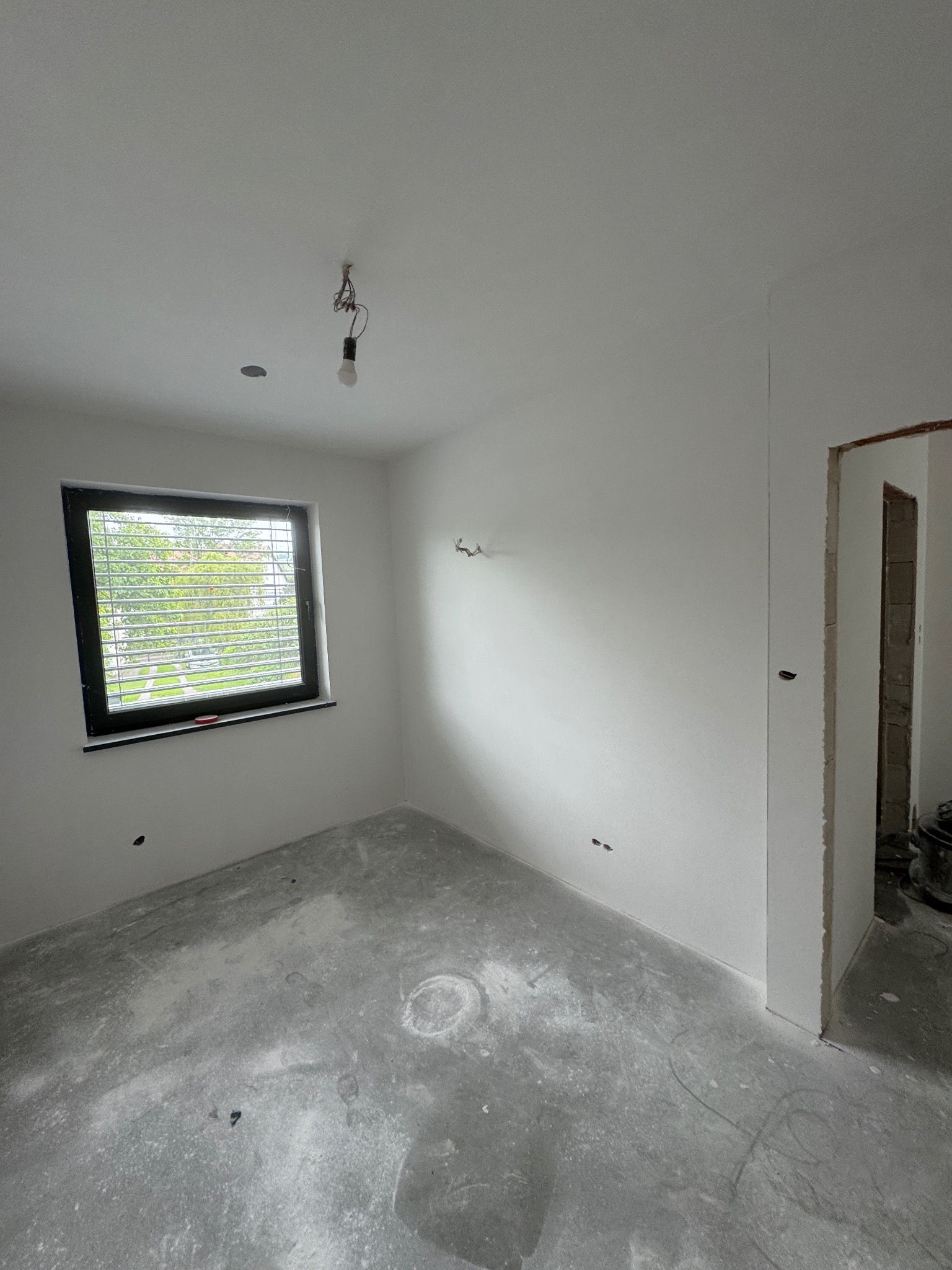 Białe ściany w pokoju po gładziowaniu, z oknem z żaluzjami i surowym betonowym podłożem. Widoczny otwór drzwiowy i wisząca żarówka.