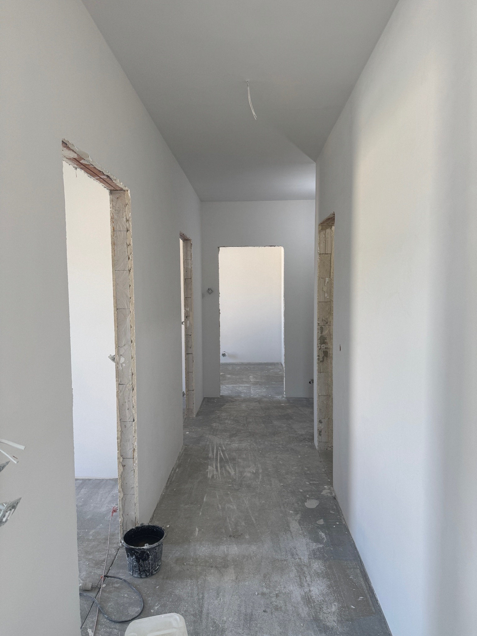 Korytarz po gładziowaniu ścian w Pietrzykowicach. Widoczne surowe ościeżnice, ściany pomalowane na biało, podłoga zabezpieczona folią. W tle widoczne kolejne pomieszczenia.