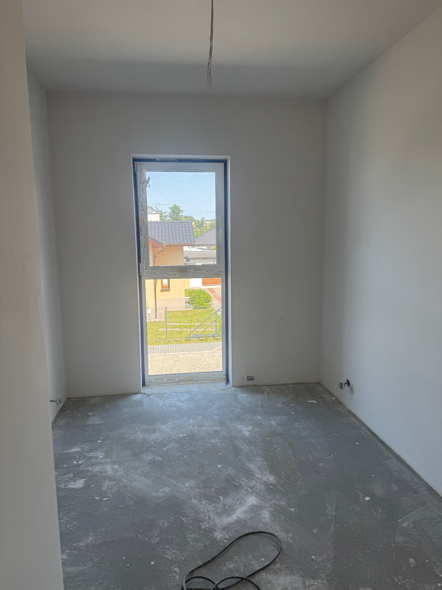 Wnętrze pokoju po gładziowaniu ścian, z widokiem na zewnątrz przez okno. Szare wykończenie podłogi i wystające kable. Delikatne światło dzienne.