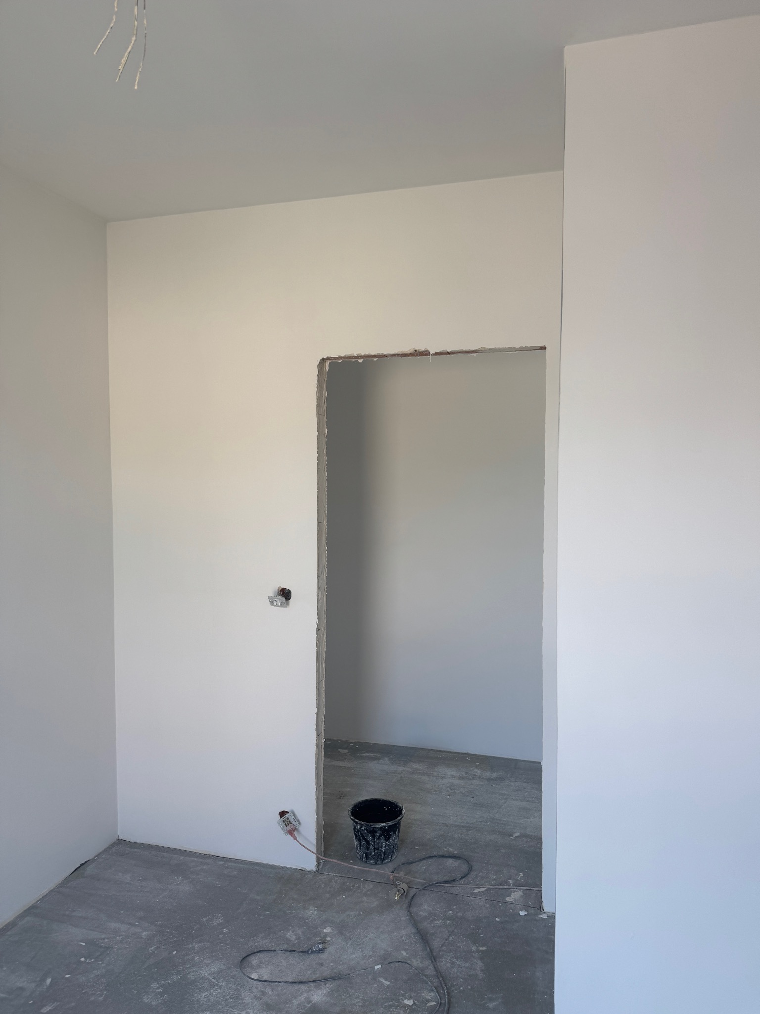 Wnętrze pokoju po gładziowaniu ścian, widoczny otwór drzwiowy bez ościeżnicy, szpachlowane gniazdko elektryczne i wiadro na podłodze.