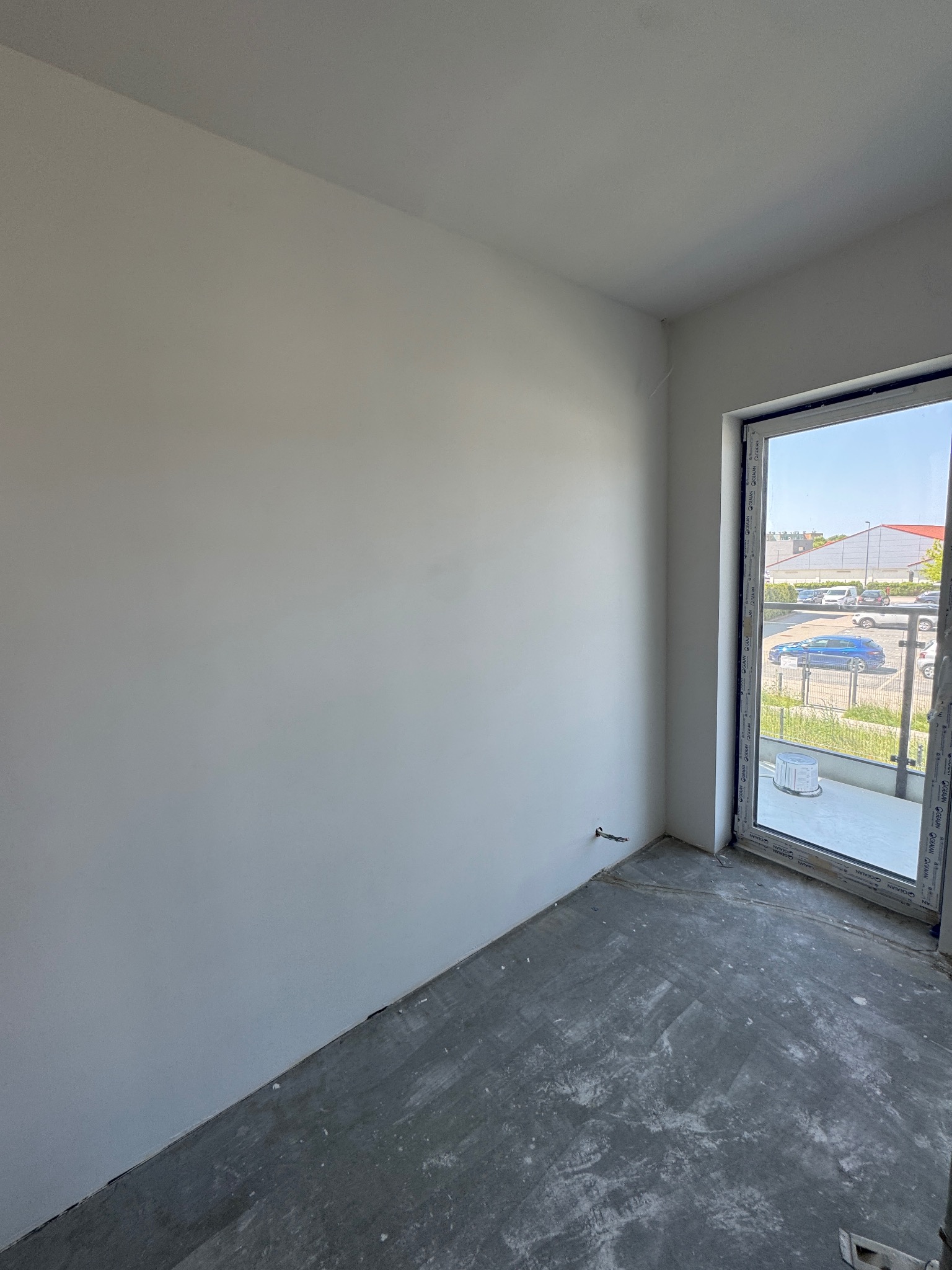 Pusty pokój z gładkimi, białymi ścianami i surową podłogą. Widoczne okno balkonowe z widokiem na zewnątrz. Minimalistyczne wnętrze w trakcie wykańczania.