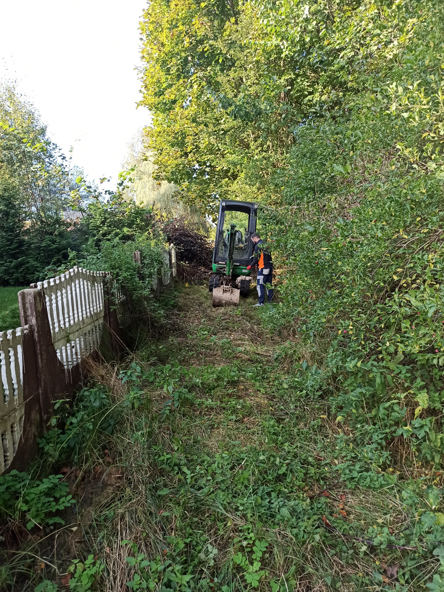 Mini koparka usuwa zarośla na działce, widoczny pracownik w odblaskowej kamizelce i fragment starego, drewnianego płotu.