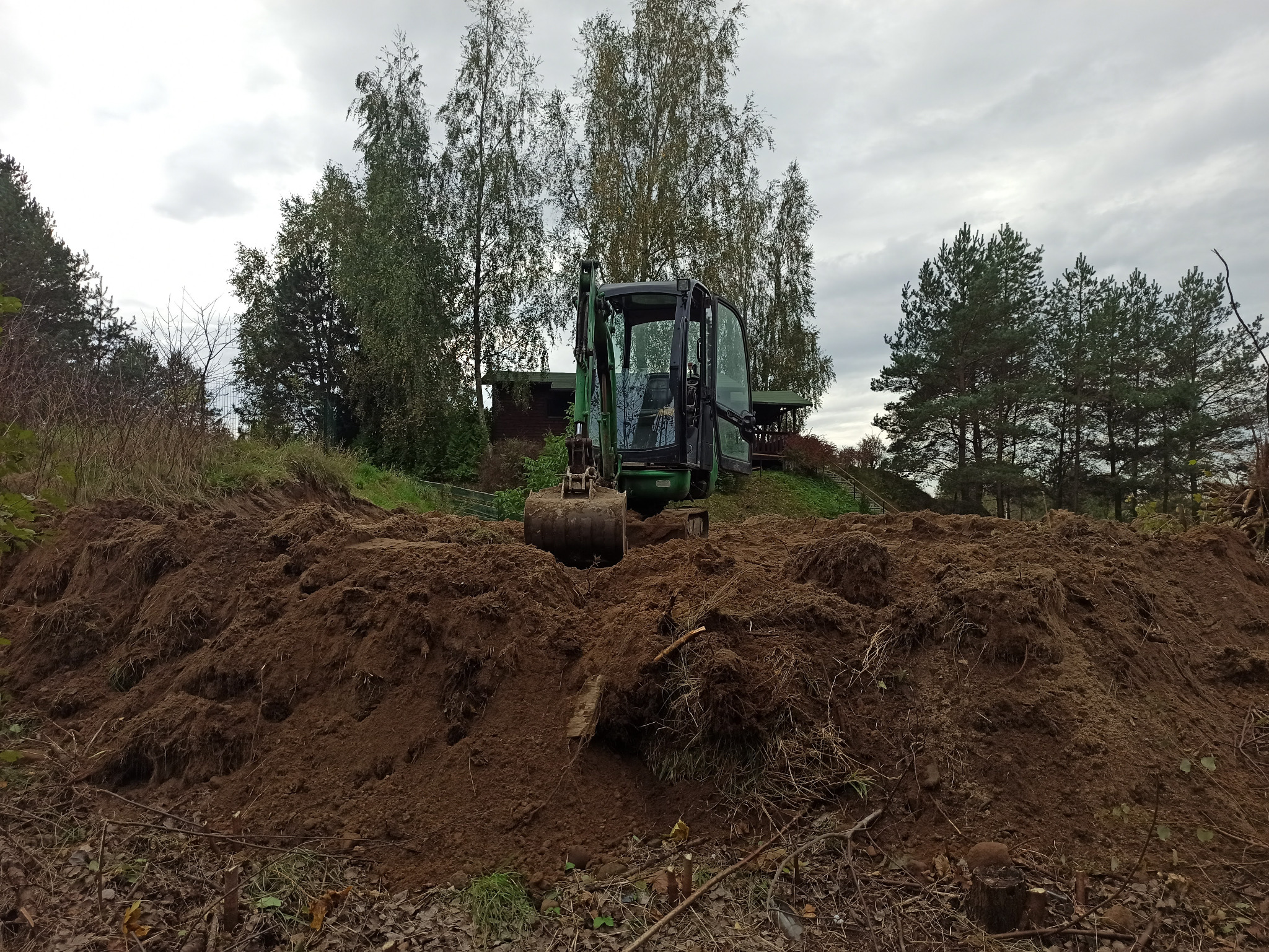 Mała koparka na wzgórzu usypanej ziemi, w tle drzewa i budynek gospodarczy.