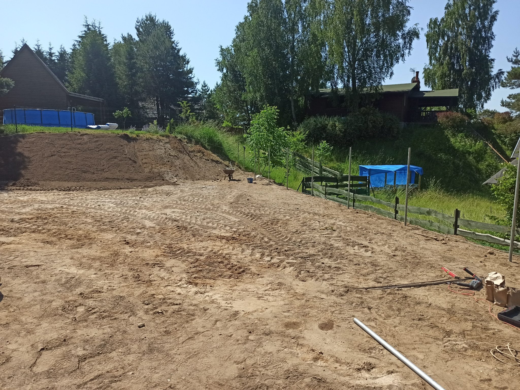 Teren przygotowany pod przyszły ogród z widocznymi śladami opon, narzędziami i elementami ogrodzenia w tle, dom z basenem w oddali.