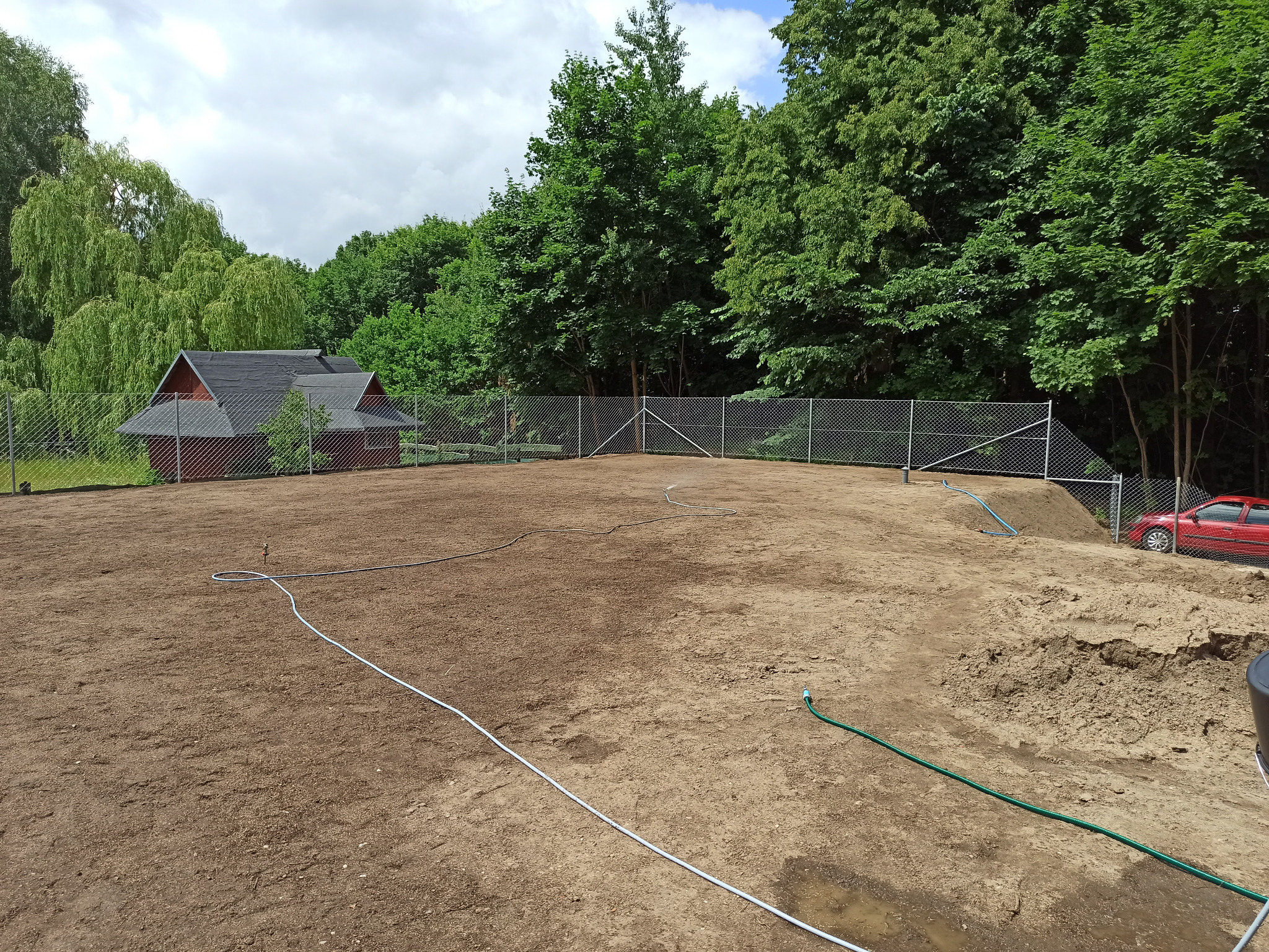 Przygotowany teren pod ogród z widocznym ogrodzeniem z siatki, budynkiem w tle i systemem nawadniającym z wężami i zraszaczami.