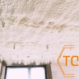 Poddasze po ociepleniu pianką, widoczne metalowe kotwy mocujące, ściany z białego betonu komórkowego, okno, logo firmy w prawym dolnym rogu.