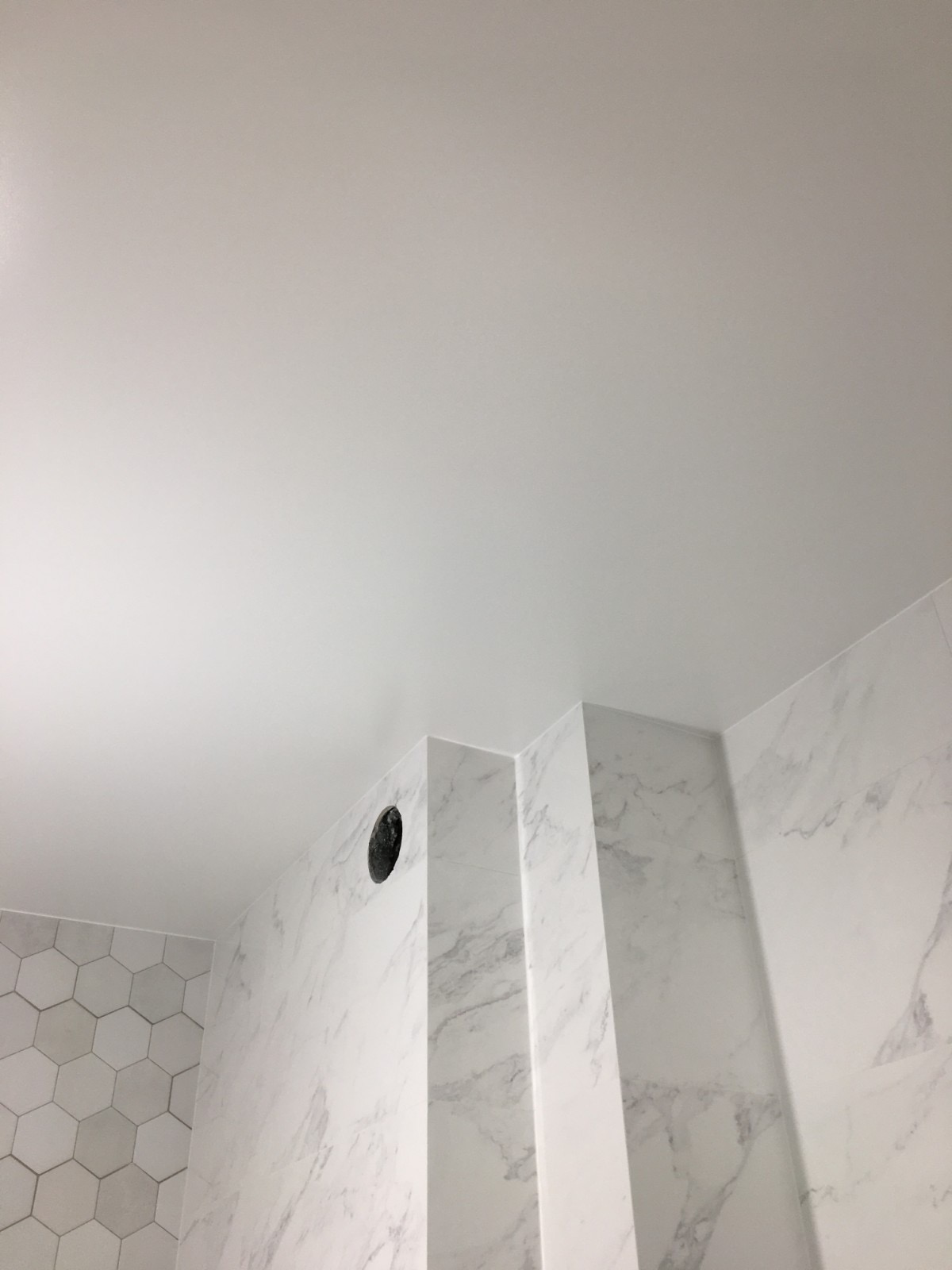 Biały sufit napinany z widocznym otworem wentylacyjnym w rogu łazienki wyłożonej marmurowymi płytkami i heksagonalnymi płytkami na ścianie.