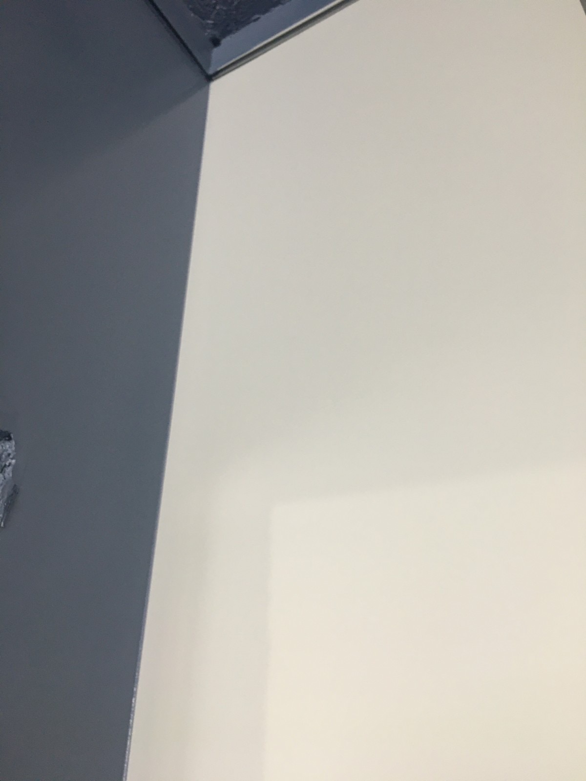 Świeżo pomalowany narożnik pokoju z widoczną linią odcięcia między szarą i kremową ścianą, z surowym, niezabezpieczonym fragmentem sufitu w rogu.