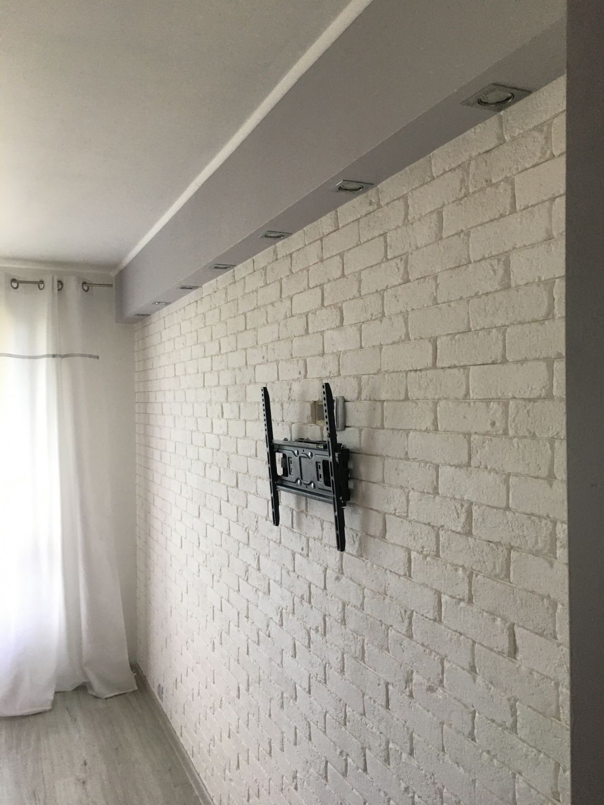 Biała ściana z cegły dekoracyjnej, oświetlenie sufitowe punktowe i zamontowany uchwyt do telewizora w jasnym wnętrzu.