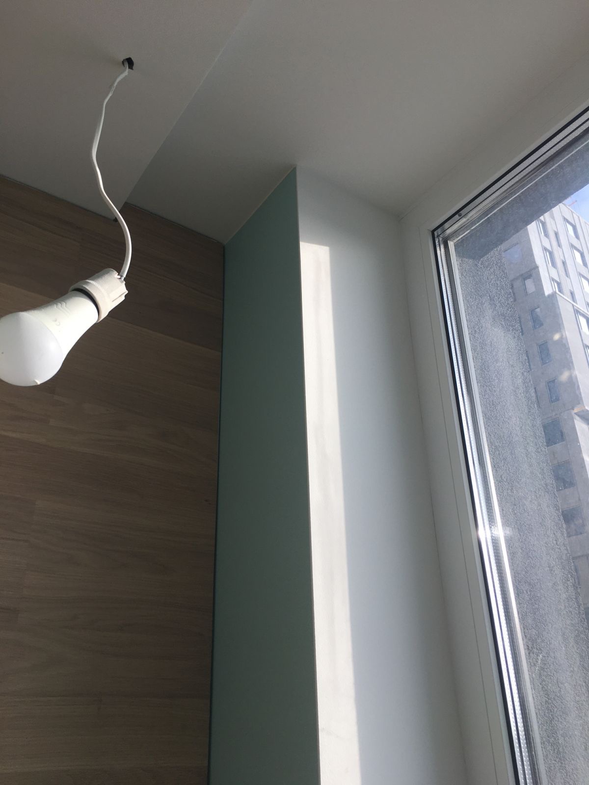 Wnętrze w trakcie prac wykończeniowych: surowa żarówka zwisająca z sufitu, fragment ściany z panelami imitującymi drewno, świeżo pomalowany na biało narożnik i okno z widokiem na budynek mieszkalny.