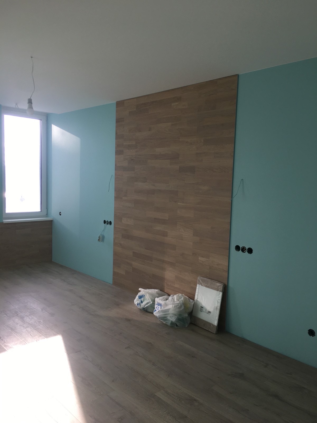 Wnętrze w trakcie wykańczania: ściana z drewnopodobnych paneli, turkusowa ściana z puszkami elektrycznymi, podłoga laminowana, worki z materiałami budowlanymi przy ścianie.