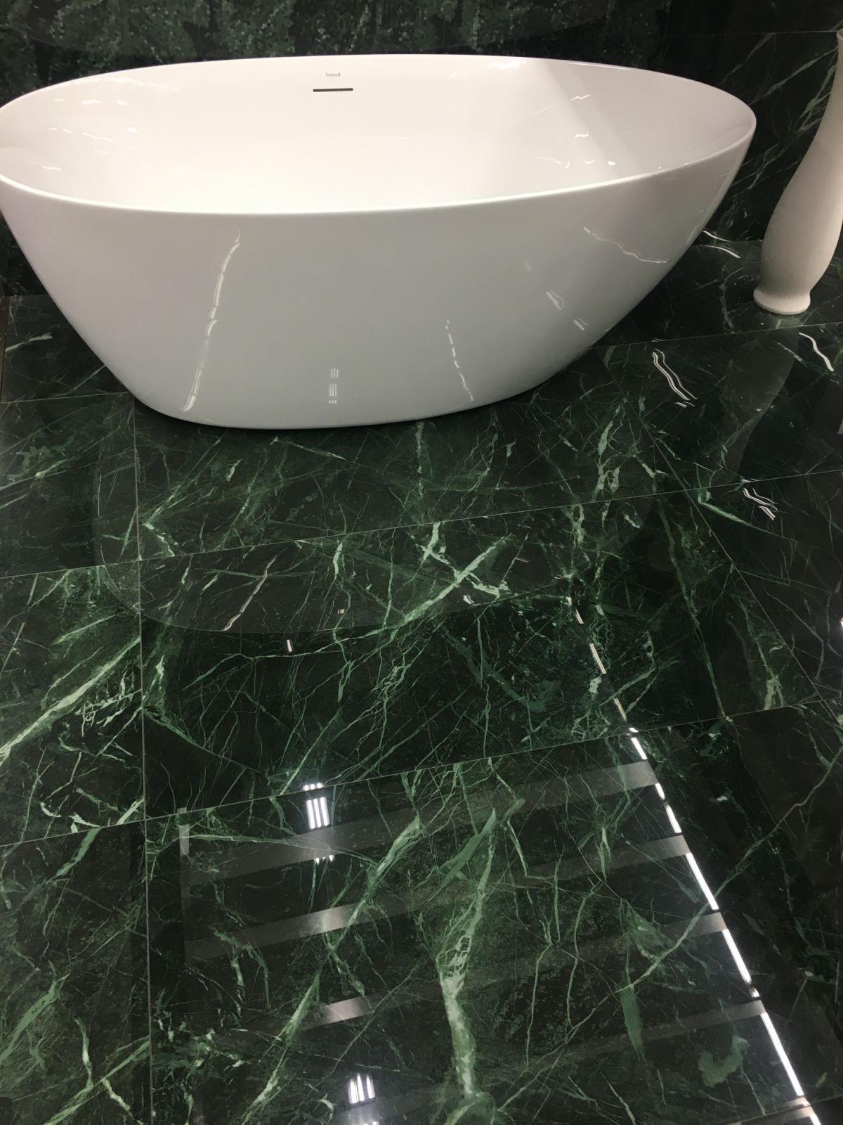 Nowoczesna, wolnostojąca wanna z armaturą na podłodze wyłożonej ciemnozielonymi płytkami imitującymi marmur z wyraźnym połyskiem.
