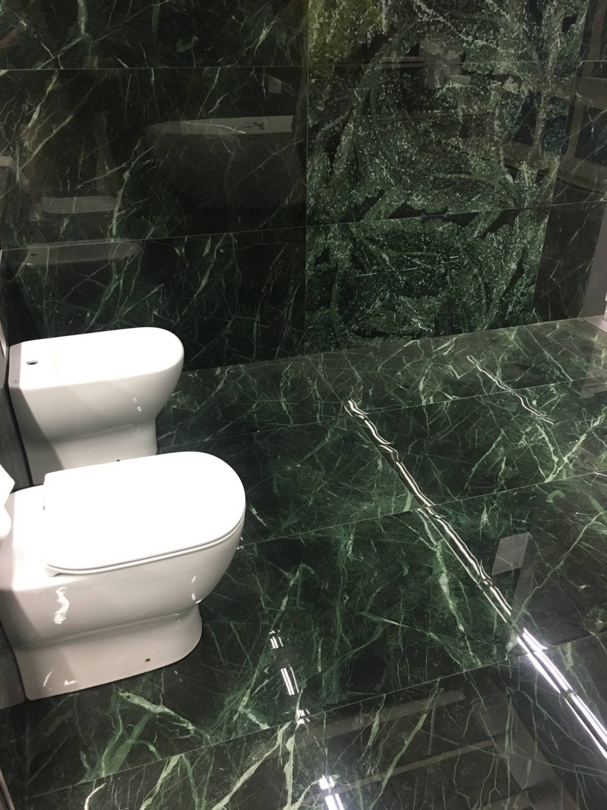 Wykończona łazienka z zielonym marmurem na podłodze i ścianach, widoczne białe sedesy.
