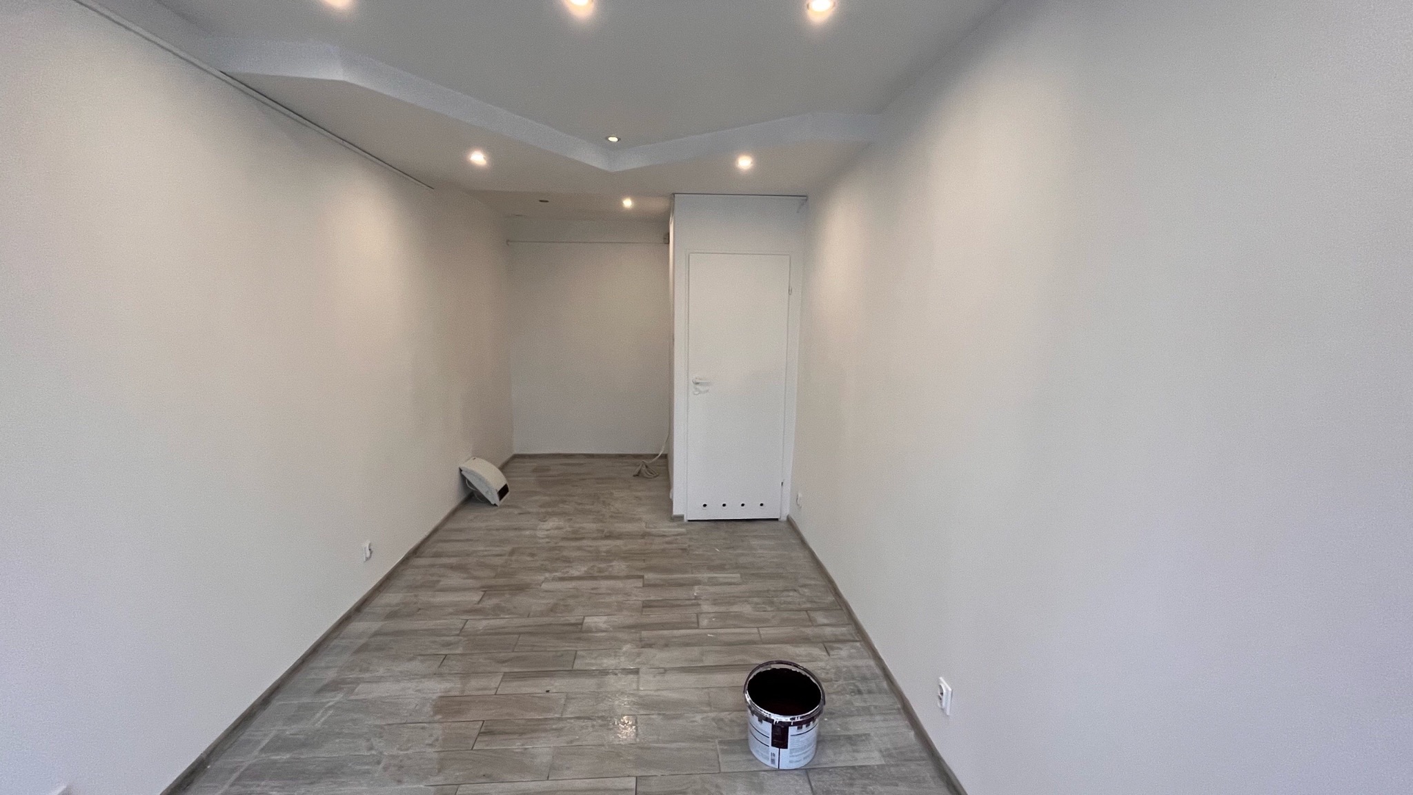 Pomalowane na biało wnętrze z ułożoną podłogą z płytek imitujących drewno, widoczna puszka z farbą na podłodze i białe drzwi w głębi pomieszczenia.