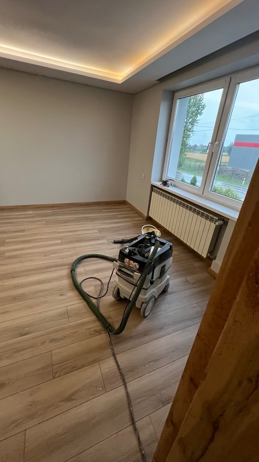 Wnętrze pokoju po remoncie z panelami podłogowymi, widoczny odkurzacz przemysłowy Festool i listwa LED pod sufitem.