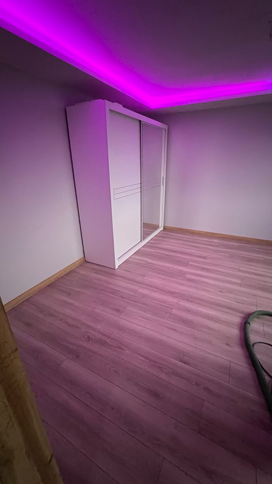 Biała szafa przesuwna z lustrami, ustawiona w pokoju z panelami podłogowymi, oświetlona fioletowym światłem LED z sufitu.