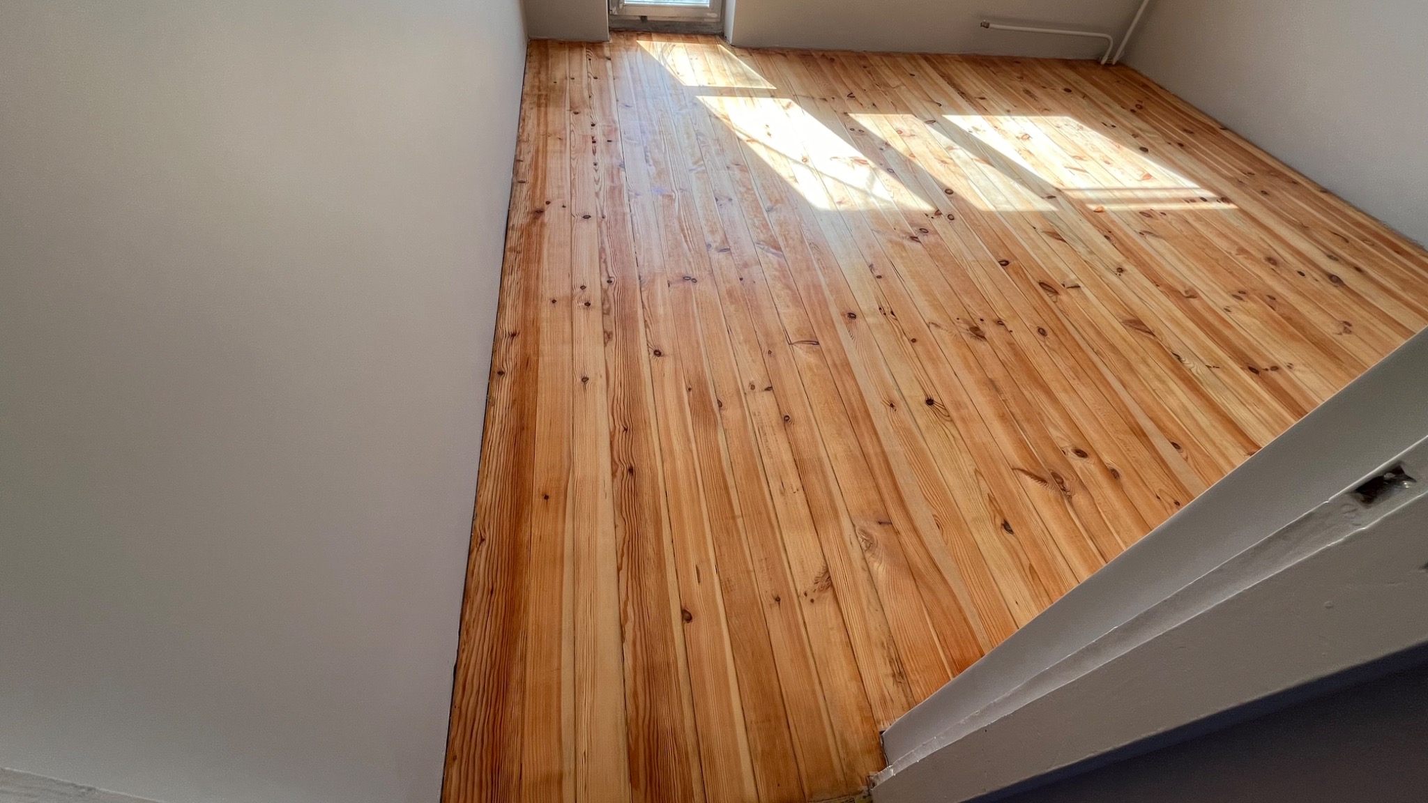 Wykończona podłoga z desek sosnowych, lakierowana na jasny odcień, widoczne promienie słoneczne wpadające przez okno, ściany w neutralnym kolorze.