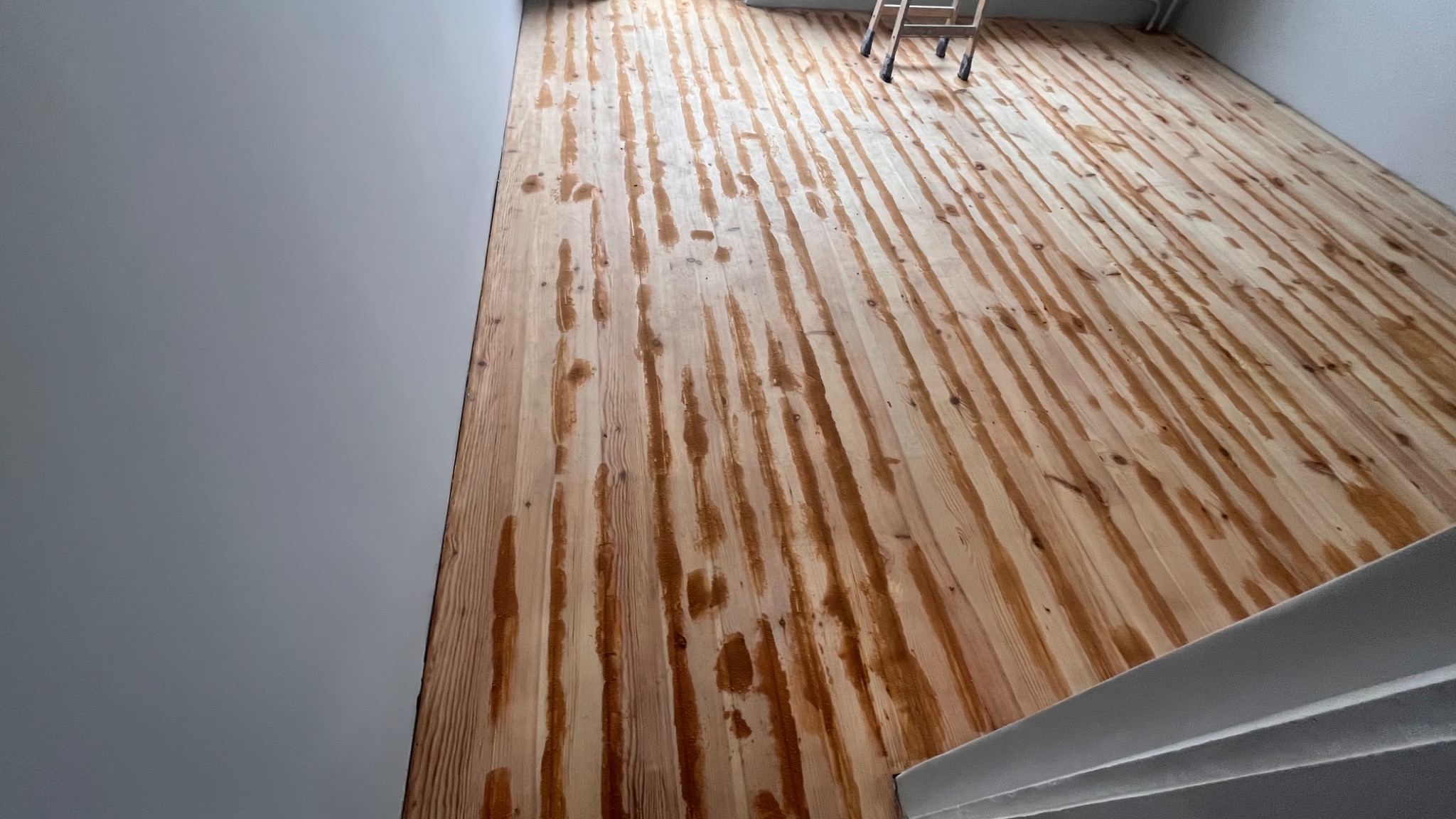 Świeżo polakierowana drewniana podłoga z widocznymi smugami lakieru, drabina w tle, jasne ściany.