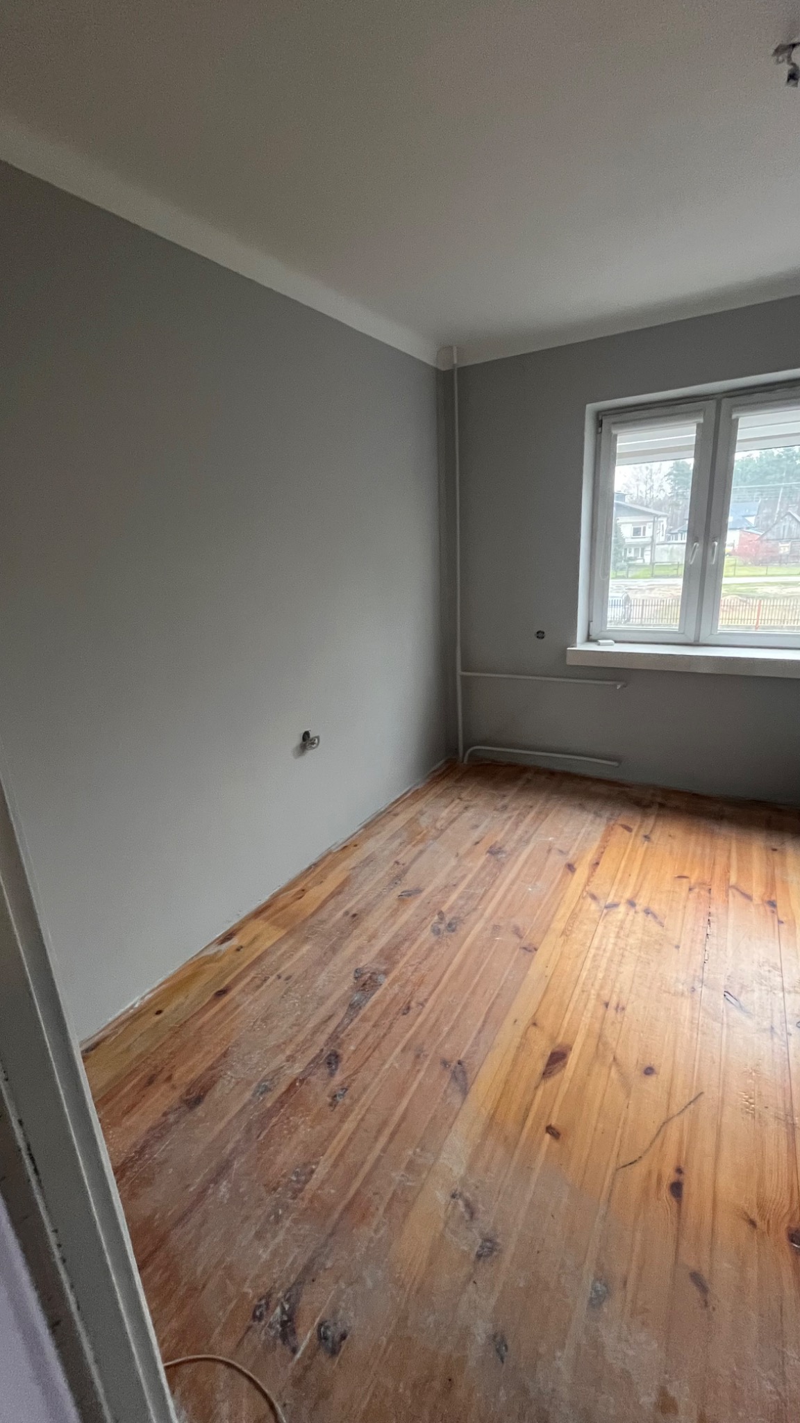 Pokój przed remontem: szare ściany z widocznymi gniazdkami, drewniana podłoga z resztkami kleju, białe listwy przypodłogowe i okno z widokiem na domy.