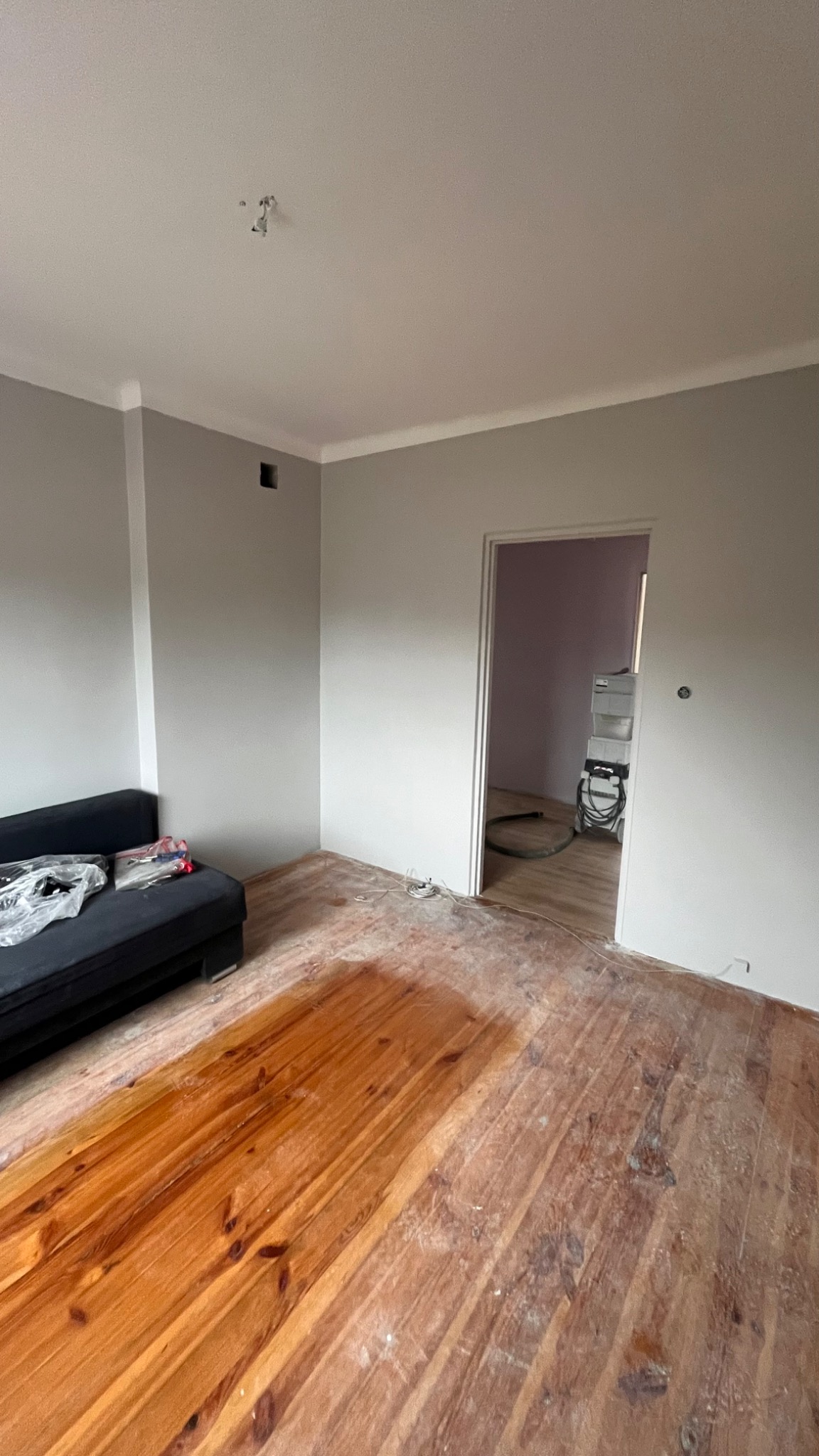 Pokój w trakcie remontu: świeżo pomalowane szare ściany z białym wykończeniem sufitu, odsłonięta drewniana podłoga, częściowo przykryta folią, czarna sofa z narzędziami, widoczny odkurzacz...