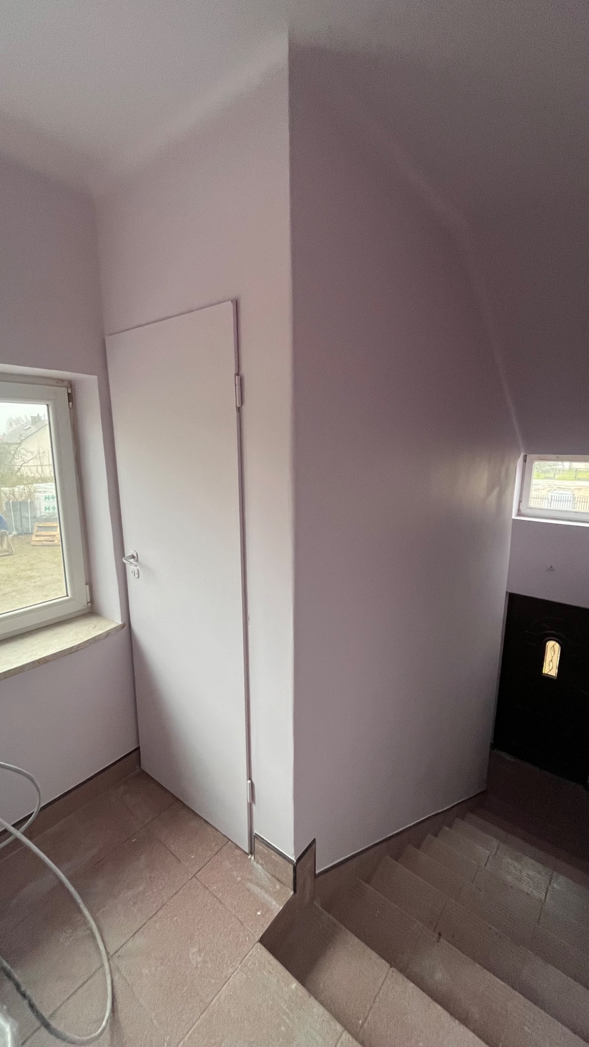 Wnętrze klatki schodowej z pomalowanymi na jasny kolor ścianami, widoczne schody z terakoty, białe drzwi wejściowe i okno z widokiem na zewnątrz.