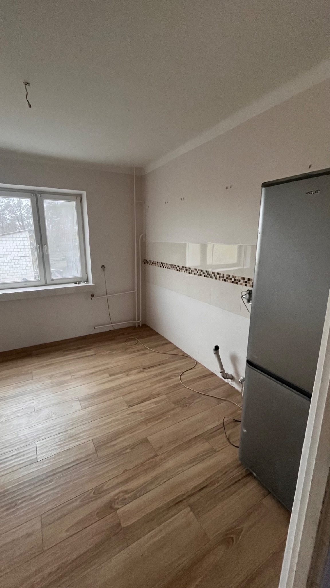 Wnętrze kuchni w trakcie remontu: widoczne nowe panele podłogowe imitujące drewno, częściowo wykończone ściany z białymi płytkami i dekoracyjnym pasem mozaiki, oraz lodówka Polar. Na suficie...